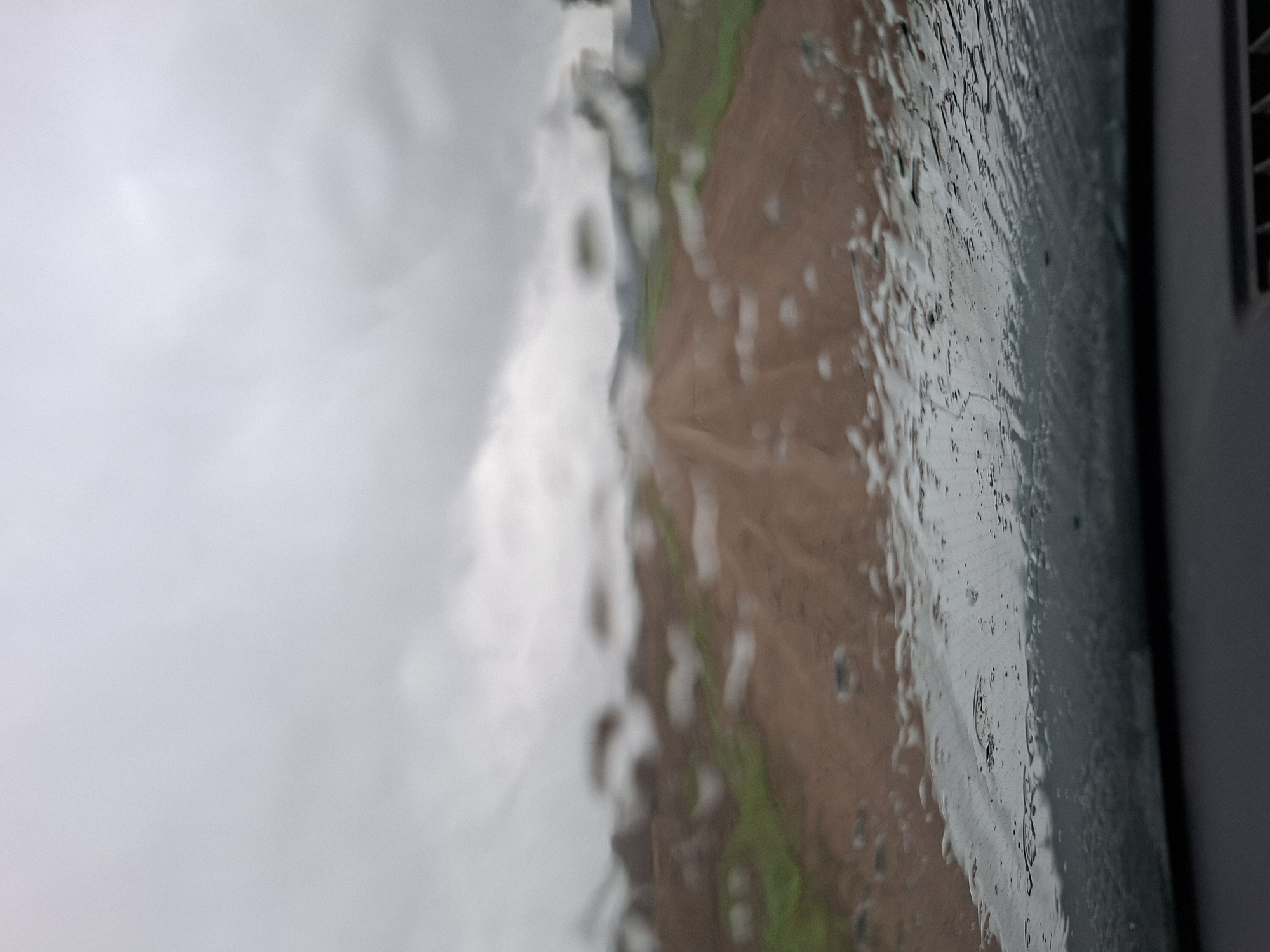 The image depicts a blurred view through a rain-streaked car windshield. Outside the vehicle, a dirt road stretches into the distance, flanked by grassy verges and what appear to be low hills or fields. The sky is overcast with gray clouds, suggesting inclement weather. The foreground is dominated by water droplets and streaks on the windshield, distorting the view of the road and surrounding landscape. The scene is devoid of any people or specific identifiable landmarks that would pinpoint a precise location within Славенскі сельскі Савет, Belarus, beyond the general impression of a rural, unpaved road under a cloudy sky. The interior of the car is partially visible at the bottom, showing a dark dashboard and a vent with horizontal slats.