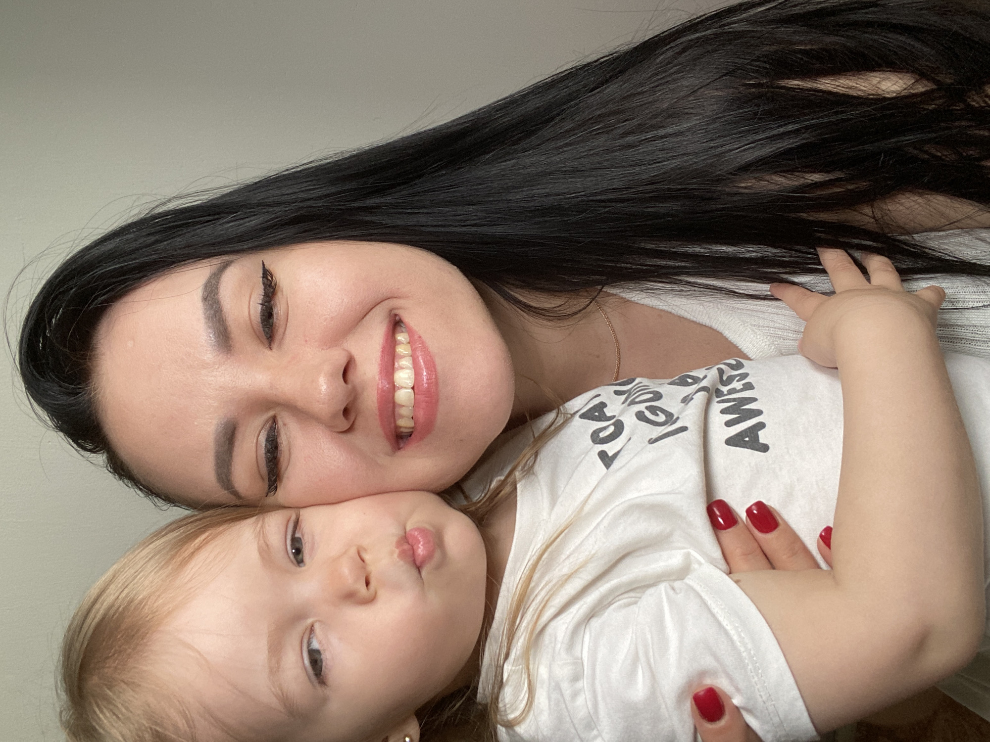 This image captures a joyful and tender moment between a mother and her young child, likely a daughter, in Chaikovsky, Russia. The photo, taken at an angle, shows the two subjects in a close, affectionate pose against a plain, light gray background, suggesting an indoor setting.
The woman, at the top of the frame, has long, straight dark hair that cascades around her and the child. She has fair skin, dark eyebrows, and a radiant, wide smile, revealing her upper teeth. She wears a light-colored, possibly white, ribbed top and a delicate gold chain necklace. Her fingernails are painted with bright red polish. Her expression conveys warmth and happiness.
Cuddled closely beside the woman's cheek is the young child, a toddler with blonde hair and fair skin. The child's lips are puckered in a playful "kissing" or "pouty" expression, looking towards the viewer. The child wears a white t-shirt, on which partial text is visible: "...AWES..." and "...SOME", likely spelling "AWESOME". A small hand, presumably the child's, gently touches the woman's hair and later rests on her chest.
The overall mood of the image is one of love, happiness, and closeness, beautifully capturing a maternal bond.