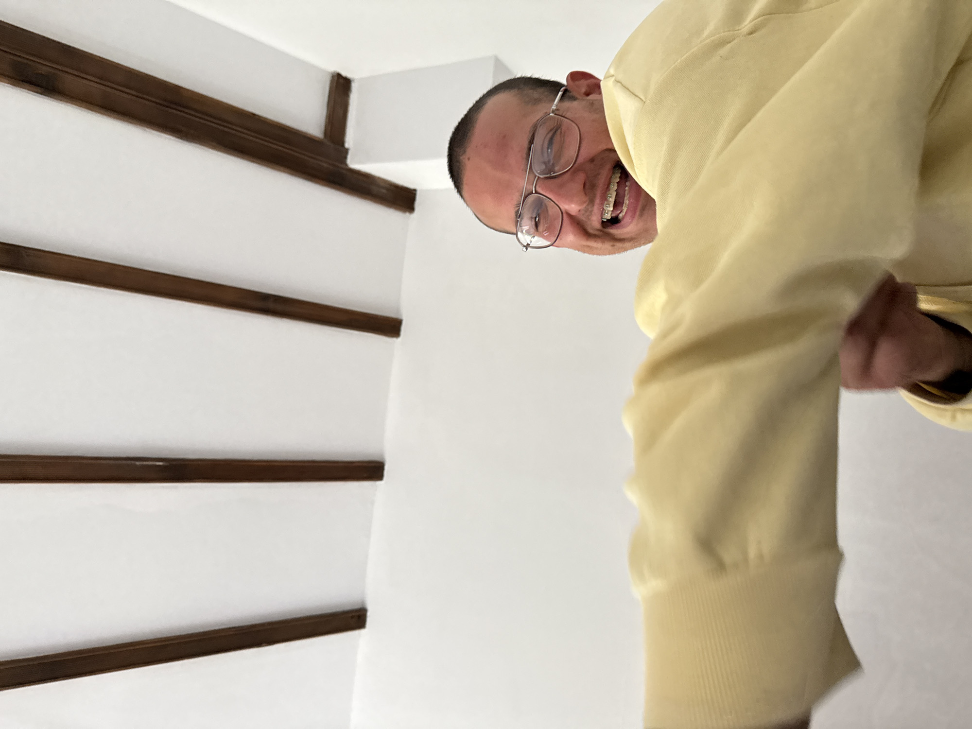 A man wearing glasses and braces is smiling at the camera, positioned in what appears to be a stairwell with white walls and dark brown wooden trim. The location is in a building with a modern design. The man is wearing a pale yellow hoodie. The orientation of the photograph suggests the camera is looking up at him from below. There is no explicit information to suggest the image was taken in Razgrad, Bulgaria.