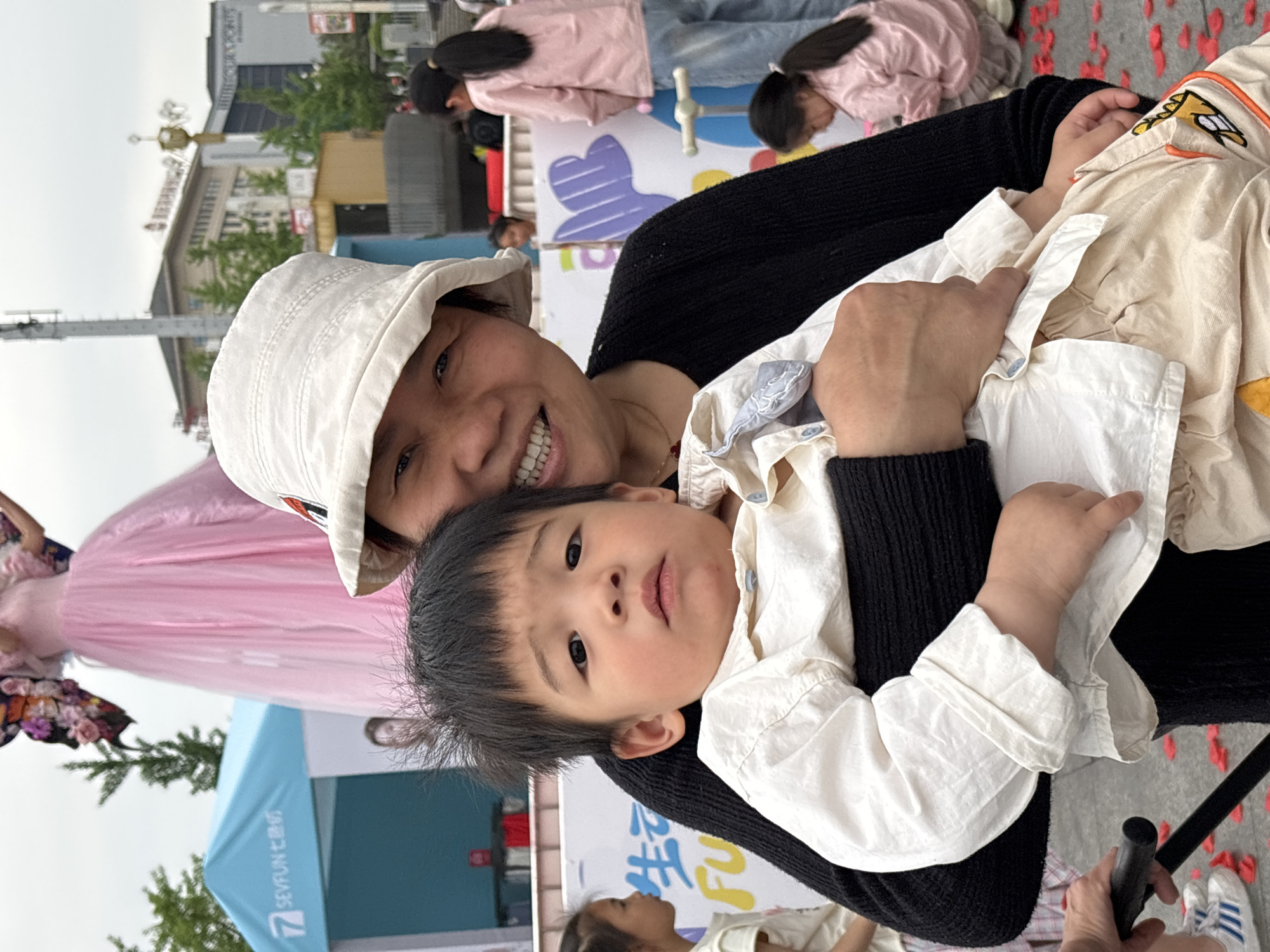 This portrait-oriented photograph captures a woman holding a young child outdoors, likely at a public event in Jiangjin, China. The woman, wearing a white bucket hat and a black long-sleeved top, smiles broadly at the camera, revealing her teeth. She holds a young child with dark hair and dark eyes, who wears a light-colored collared shirt and a light beige outer jacket. The child looks towards the camera with a neutral, curious expression.
The background indicates a lively, festive setting during the daytime, possibly under an overcast sky. To the left, a person in a long, flowing pink dress with large butterfly wings is visible near a green tree and a blue tent. The tent features the logo "SEVFUN" and the Chinese characters "七色纺" (Qī Sè Fǎng), translating to "Seven Colors Textile" or "Seven Colors Spinning." Behind the main subjects, a colorful backdrop with abstract shapes, including a large purple heart, can be seen. In the further background, urban buildings are visible, one partially displaying the text "WHITEFOUR POINTS." Scattered red petals adorn the ground in the lower right, alongside a black pole and a pair of white and blue Adidas sneakers. Other blurred figures, including children in pink jackets, populate the scene, contributing to the event atmosphere. The woman appears happy and engaged, while the child seems observant of their surroundings.