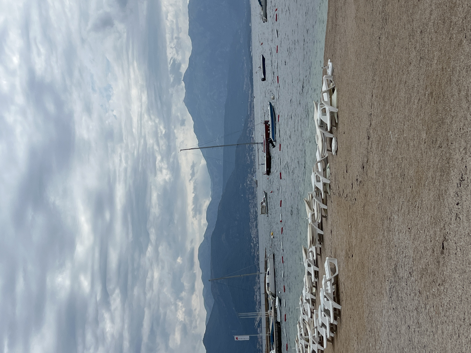 This image depicts a tranquil coastal scene in Baošići, Montenegro, characterized by an overcast sky and calm waters. In the foreground, a long line of white plastic beach chairs is neatly arranged on a light brown, possibly sandy or pebbly, shore. The chairs are empty, suggesting a quiet moment, perhaps early morning, late afternoon, or an off-peak season.
Beyond the shoreline, the calm, light blue-grey water stretches out, dotted with several boats. A prominent sailboat with a tall mast is visible, alongside smaller motorboats. Some boats appear to be anchored or gently floating. On one of the closer boats, a red and white flag or banner can be seen, with the word "SAIL" partially legible.
In the midground, impressive mountains rise steeply from the water's edge, forming a dramatic backdrop. These mountains are covered in deep blue-grey hues, suggesting distance and their imposing scale. The peaks of the mountains are partially obscured by the extensive cloud cover that dominates the sky. The sky is entirely overcast with thick, light grey clouds, creating soft, diffused lighting across the entire scene. There are no people visible, contributing to the peaceful and serene atmosphere of this coastal landscape.