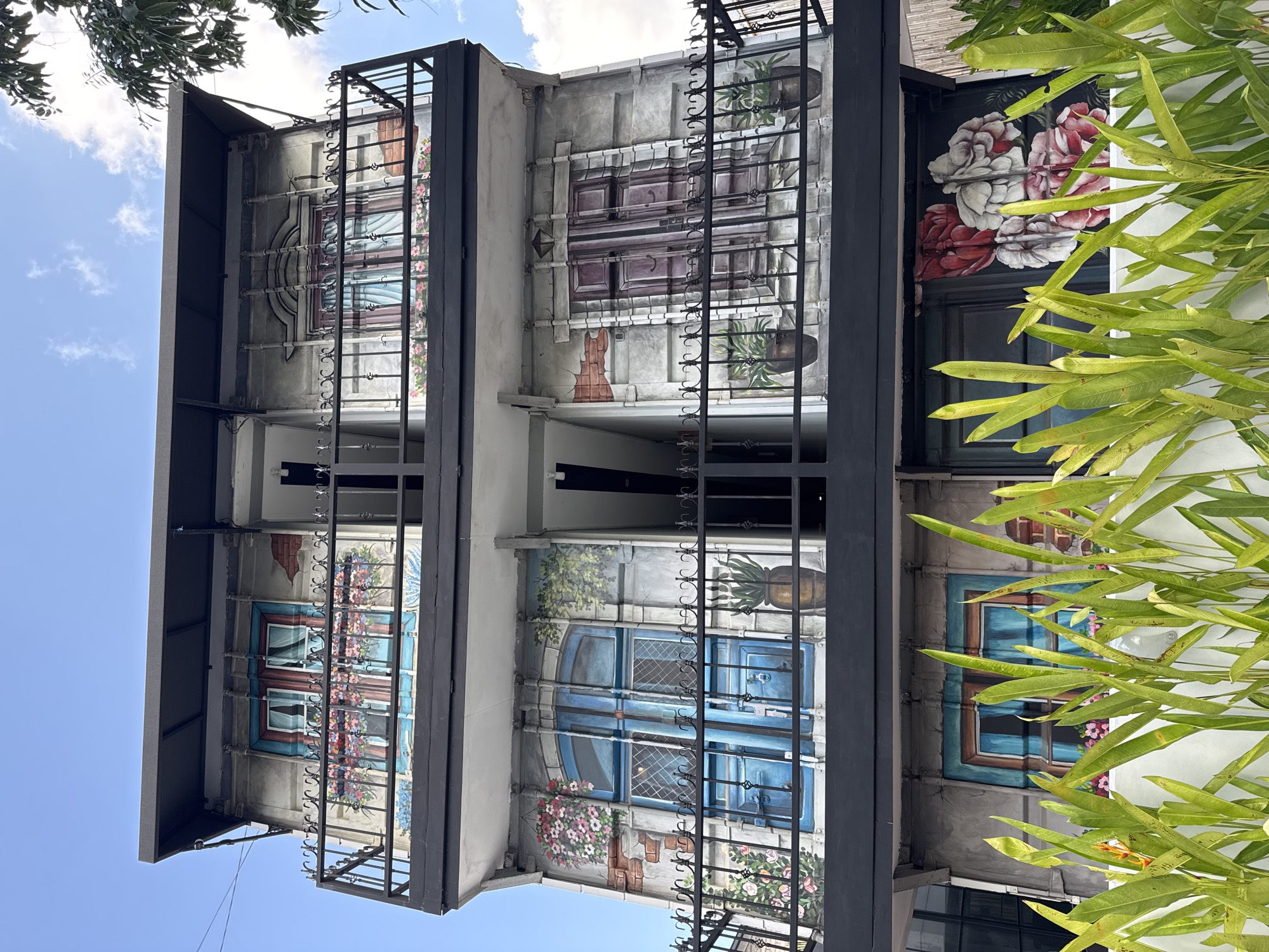 The image shows a multi-story building with an exterior decorated with murals of windows, doors, and floral arrangements, designed to resemble European architecture. The building has black metal balconies and railings on each level, and is viewed from a low angle. In the foreground, lush green plants with long, pointed leaves partially obscure the lower portion of the building. The sky above is blue with scattered white clouds. The scene is located in North Kuta, Indonesia, and appears to be a street-level view of a building façade. There are no people visible in the image.