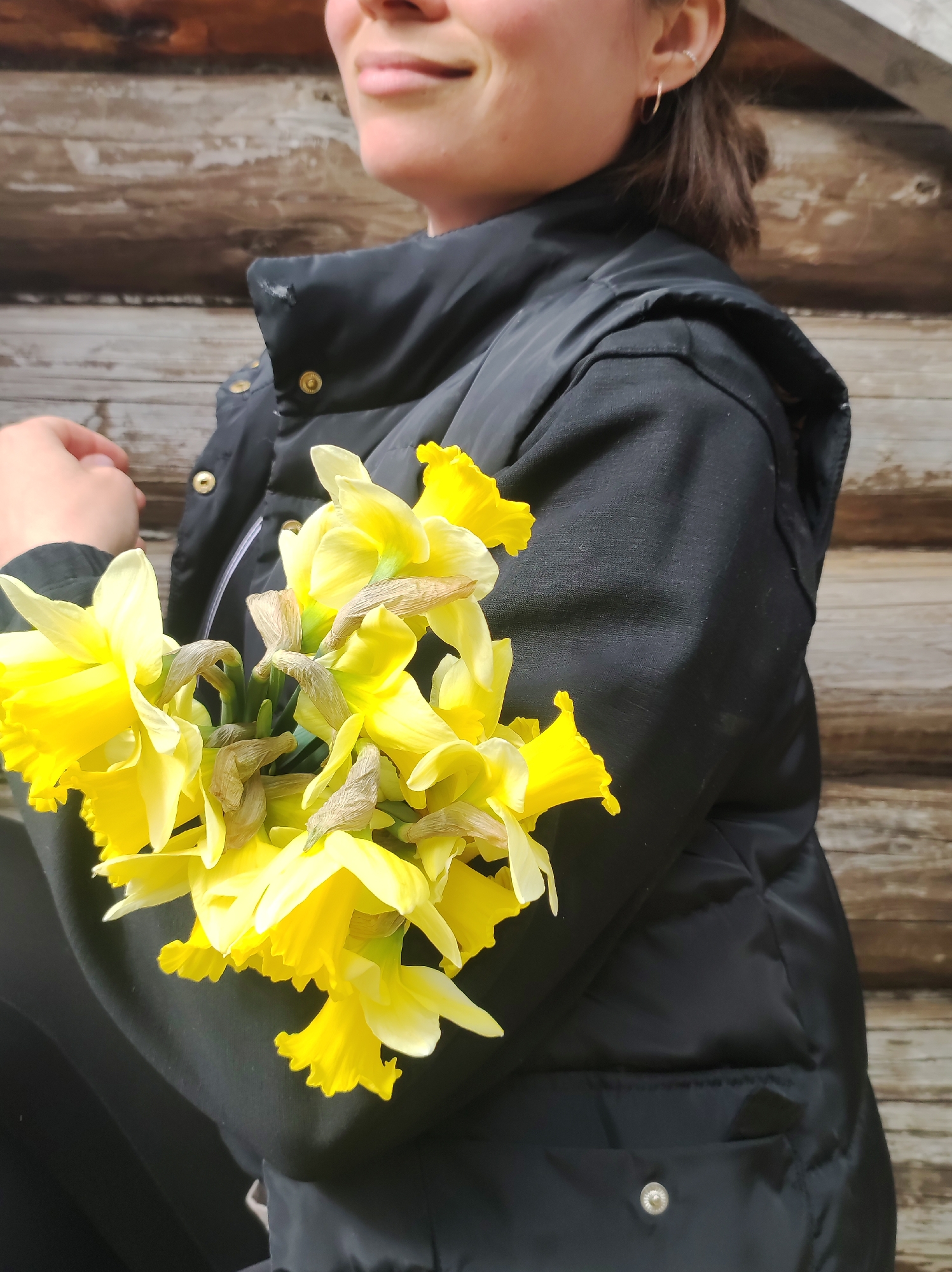 A woman stands outdoors in front of a wooden wall, holding a bouquet of yellow daffodils. The woman is wearing a black puffer jacket. She has her dark hair pulled back, and is wearing earrings. The photograph's location, Фоки, Russia, is not discernible from the visual elements.