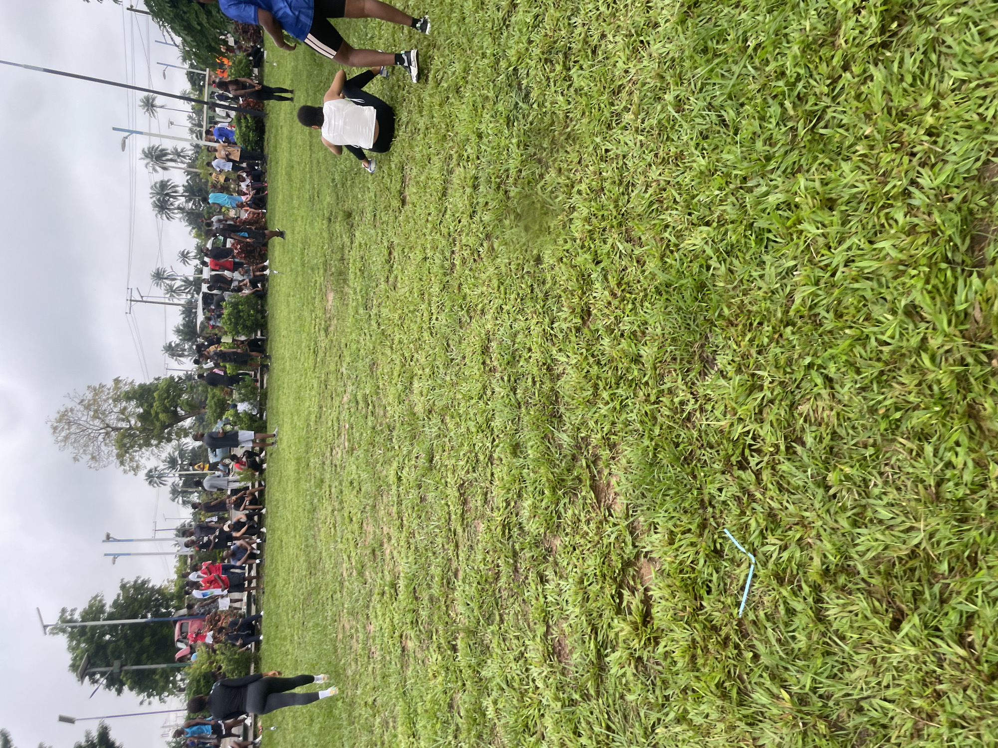 The image depicts an outdoor gathering of people in what appears to be a park or open field. The ground is covered in green grass, with some patches of bare earth. In the background, there are trees, palm trees, and some structures that resemble streetlights or utility poles. A group of people are visible in the middle ground, some standing and others sitting on the grass. They are dressed in casual attire, with many wearing athletic clothing. Several individuals are seen walking or standing, suggesting a social event or a casual get-together. The sky is overcast, indicating a cloudy day. The presence of many people in an open, grassy area, along with the casual attire, suggests a community event, a sporting activity, or a social gathering in Ihiagwa, Nigeria.