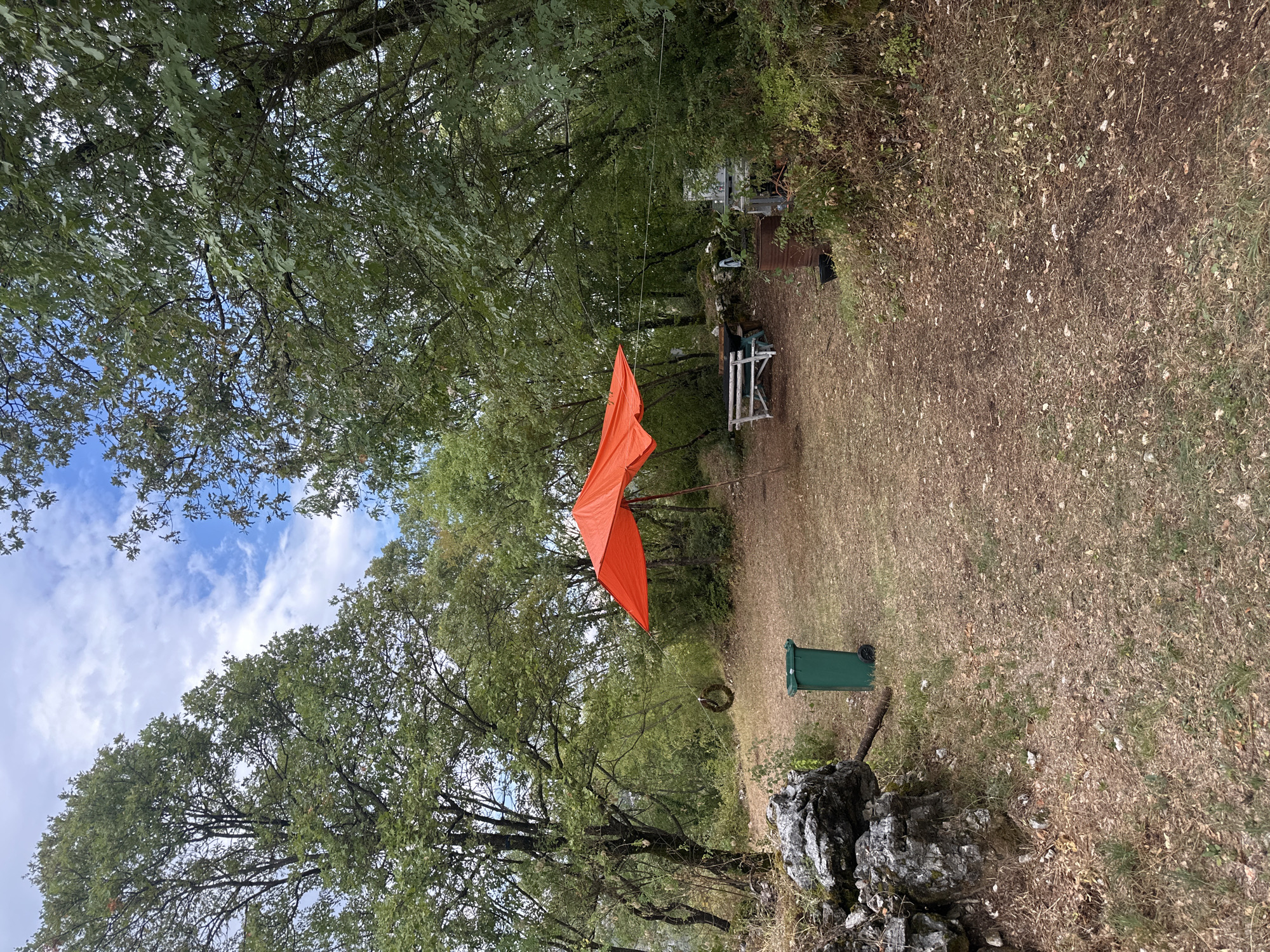 The image depicts an outdoor scene in a wooded area, possibly a campsite or picnic spot, located in the vicinity of Cetinje, Montenegro. In the foreground, to the left, a pile of large grey rocks is visible, with dry grass and fallen leaves covering the ground. A green trash can with wheels stands near the rocks. Further into the scene, an orange tarp is stretched over a makeshift frame, providing shade. Beneath the tarp, a white wooden bench and a dark-colored table are placed on the dry, grassy ground. A circular wreath hangs from a rope tied to a tree on the left side of the image. In the background, dense green foliage of trees and bushes dominates the landscape, with a blue sky with scattered white clouds visible above. To the right of the bench, a white table-like structure and some containers are partially obscured by vegetation. The overall impression is a rustic, natural setting.