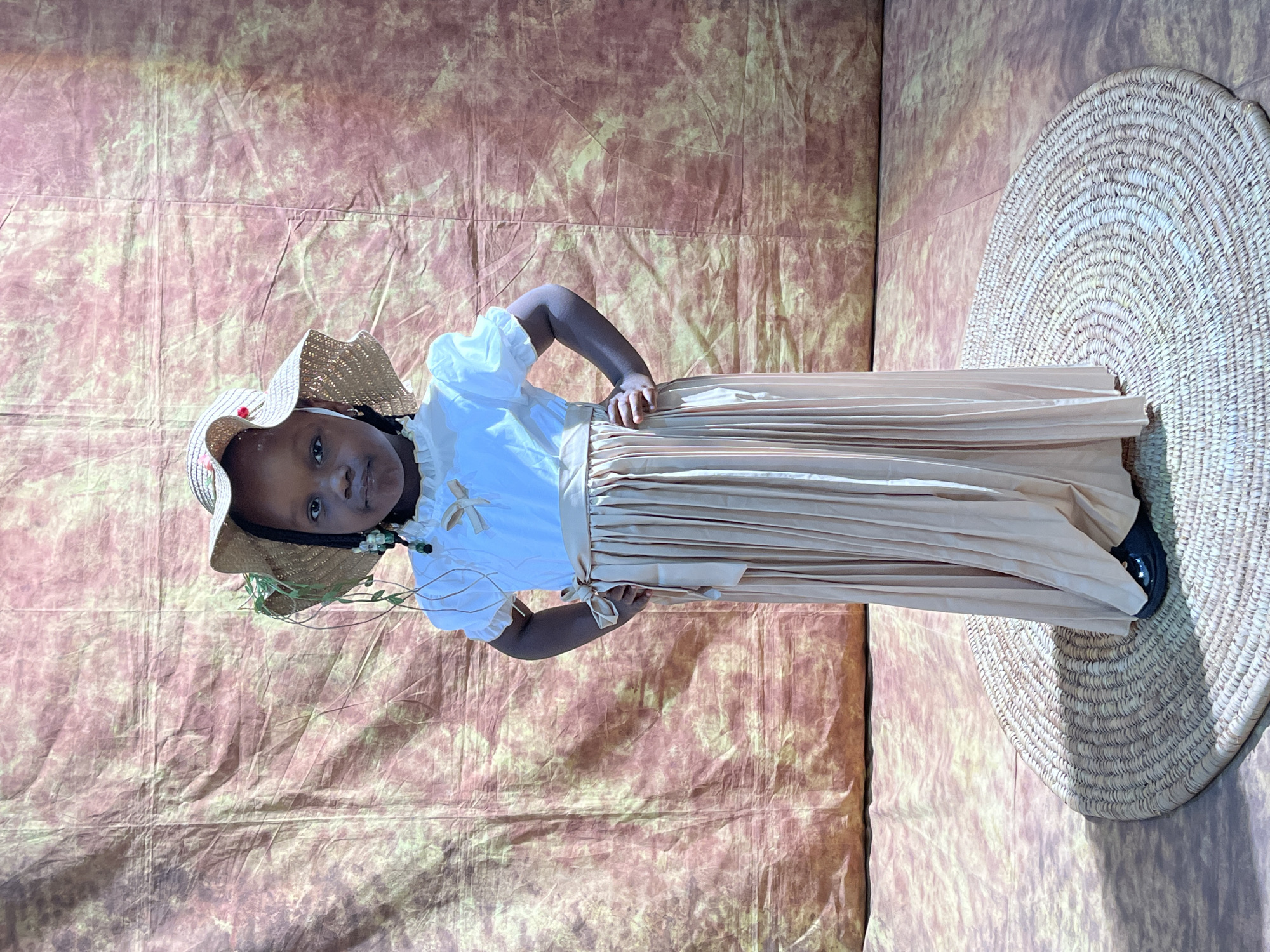 A young Black girl stands center frame wearing a white puffed-sleeve blouse, a beige pleated skirt, and a straw hat adorned with greenery and small red flowers. She has dark skin, dark eyes, and her dark hair is braided and decorated with beads. Her hands are on her hips. She is standing on a circular woven mat in front of a textured backdrop with a gradient of pink and yellow hues. The setting is indoors, possibly a photo studio, and no specific location in Barakin Akawo, Nigeria can be determined from the image alone.