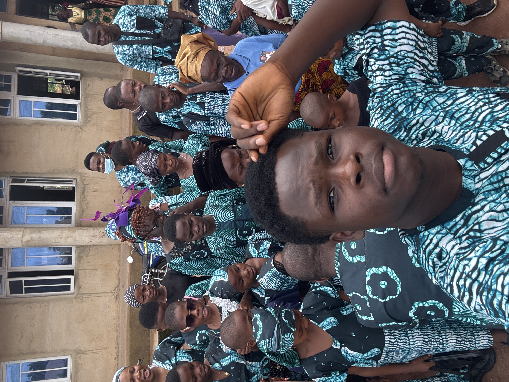 A gathering of people, mostly adults and some children, is captured in Eziudo, Nigeria. Many are clad in similar patterned shirts, predominantly in shades of black, blue, and white. A few wear different attire. In the foreground, a man's face is prominent, with a hand partially obscuring the view above his head. Behind him, a crowd extends, some looking towards the camera while others are engaged in different activities. The background reveals a building with multiple windows, suggesting an outdoor setting adjacent to a structure. A motorcycle is also visible amidst the gathering.