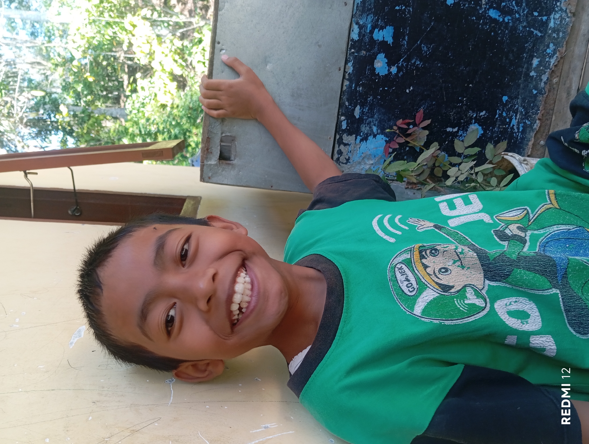 A young boy with short, dark hair and a wide smile is pictured outdoors, leaning against a light-colored wall. He is wearing a green t-shirt with a graphic print featuring a cartoon character in a helmet and the text "GOJEK". His right arm is extended, resting on a weathered, metallic surface that appears to be part of an outdoor structure, possibly a cabinet or enclosure. The background shows lush green foliage and trees, indicating a natural or semi-rural environment. A portion of a wooden window frame is visible on the left. The image appears to have been taken in Pagar Alam, Indonesia, given the presence of "REDMI 12" in the lower left corner, suggesting the phone used for the photograph.