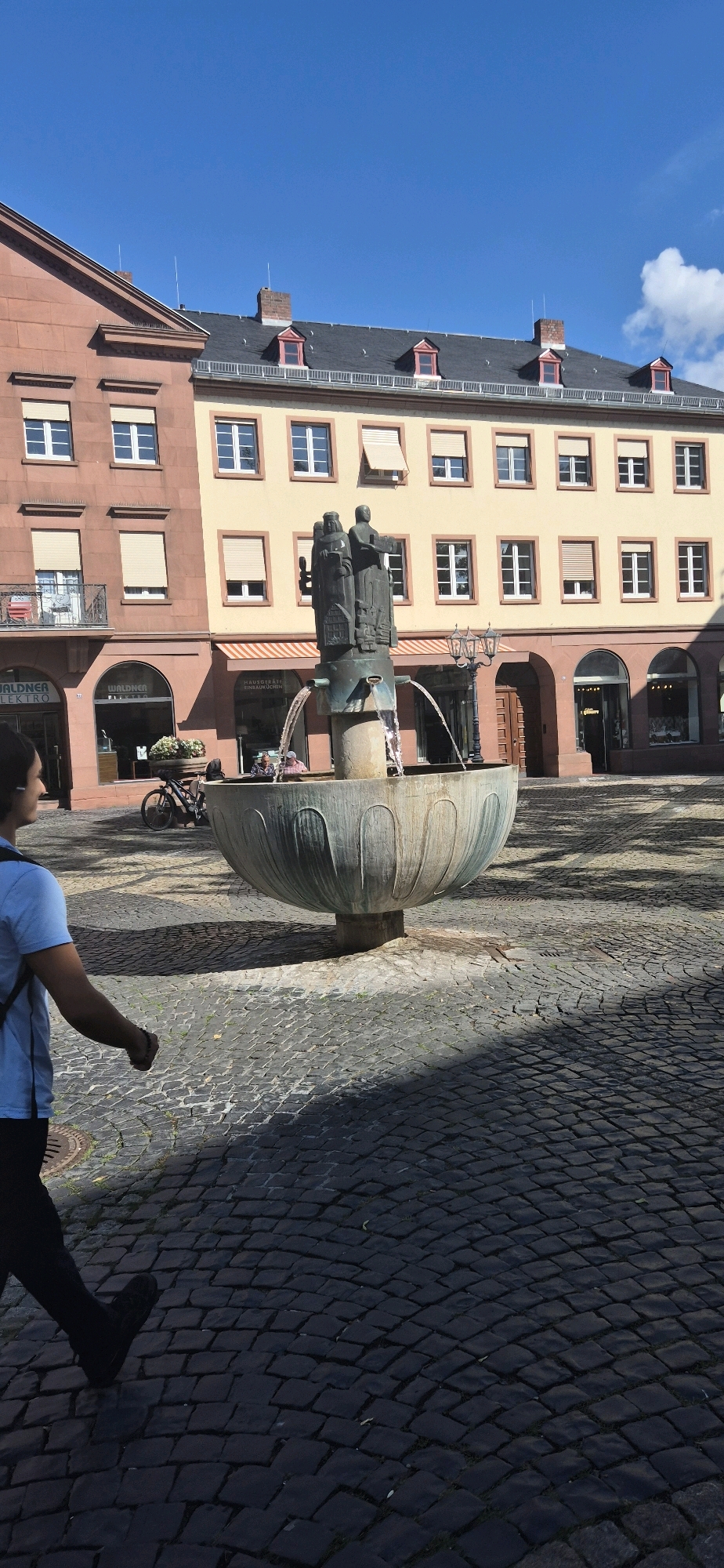The image depicts a view of the market square in Mainz, Germany, featuring a large, ornate fountain as the central element. The fountain is made of a weathered, greenish-bronze material and has a multi-tiered sculpture on top depicting several figures. Water cascades from the top tier into a wide, shallow basin. The square is paved with dark cobblestones, and shadows from nearby buildings and a street lamp are cast across the ground.
In the background, traditional European architecture lines the square. The buildings have facades of red sandstone and pale yellow plaster, with multi-paned windows and red-tiled roofs featuring dormer windows. Some of the windows are covered with beige roller blinds. Storefronts are visible at street level, including one with a sign reading "WALDNER ELEKTRO" and another with "WALDNER BISTRO." A bicycle is parked next to a planter box filled with flowers.
In the foreground, a person wearing a blue t-shirt and dark pants is walking from left to right, their back mostly to the camera. They appear to be carrying a backpack. The sunlight illuminates the scene, creating a bright, clear day with a blue sky. The overall impression is one of a historic European city square on a sunny day.
