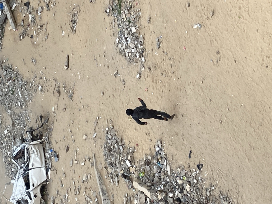 The image presents a high-angle view, likely from above, looking down onto a desolate and debris-strewn landscape. The predominant ground cover is light brown, sandy earth, which appears uneven and marked by various foot impressions or small undulations.
Centered in the frame, a single individual is visible lying prone on the sandy ground, facing downwards. The person is dressed entirely in dark clothing, possibly black, covering their head as well. Their limbs are somewhat outstretched, with one arm appearing to be slightly bent at the elbow. Their posture suggests they are motionless, possibly injured, unconscious, or deceased.
The ground around the person and across much of the frame is littered with significant amounts of rubble and debris. These include numerous jagged pieces of light grey and darker colored concrete or rock fragments, varying in size. In the bottom left corner of the image, the heavily damaged and mangled remains of a white or light-colored vehicle, possibly a car, are visible, crushed and distorted. Scattered amongst the larger rubble are smaller pieces of refuse, including what might be plastic or fabric scraps.
The lighting indicates daytime, with a clear illumination of the scene, though specific shadows are not prominent enough to determine the exact time of day. The overall scene conveys an impression of devastation or aftermath, with the motionless figure adding a somber and concerning element to the environment of widespread destruction. No text is visible in the image.