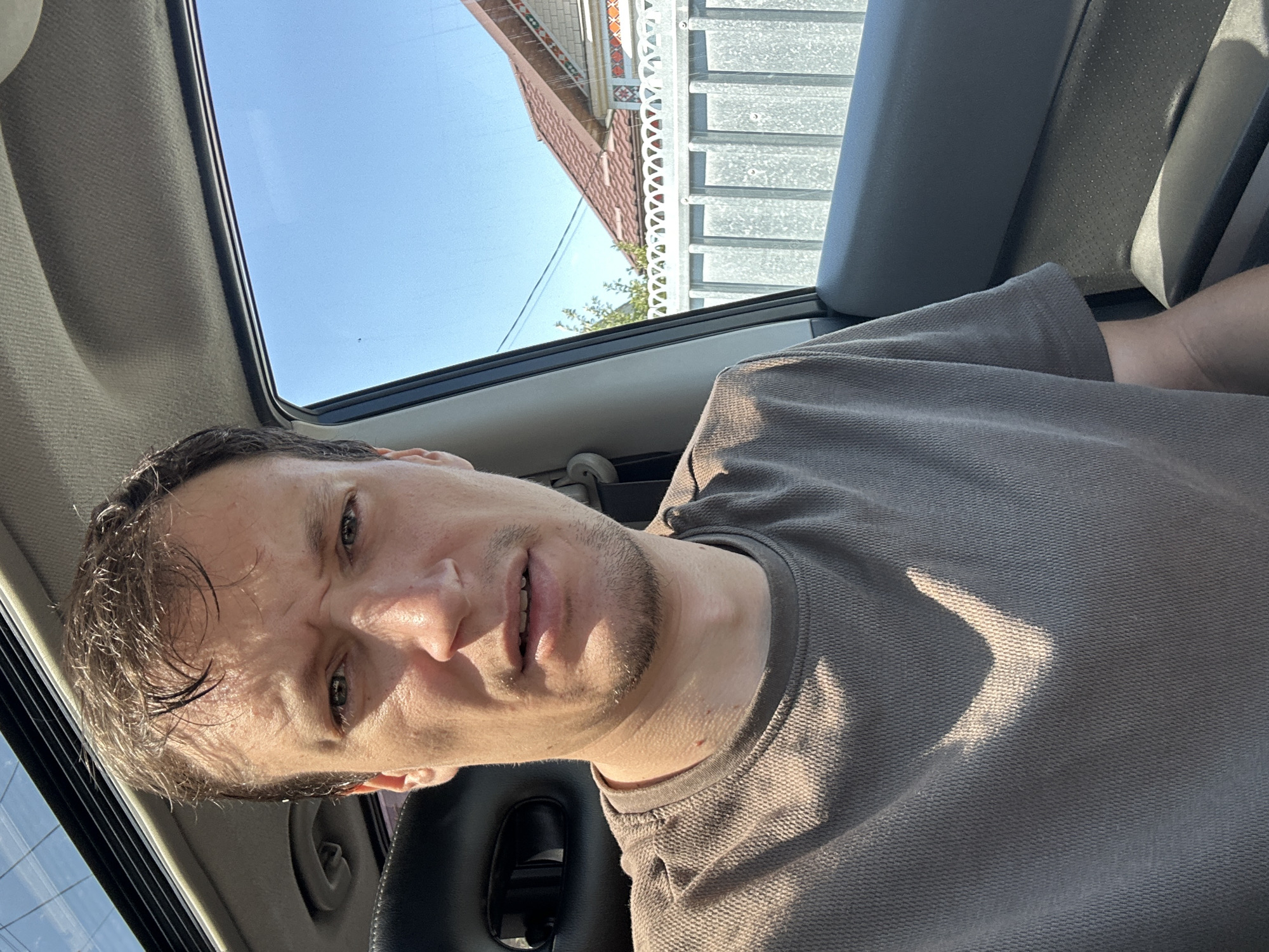 A selfie taken from inside a car shows a man with short, dark, disheveled hair, looking directly at the camera with a concerned or confused expression. His eyebrows are furrowed, and his mouth is slightly open. He is wearing a short-sleeved, textured, brown t-shirt. Sunlight is casting a shadow on his shirt, creating a V-shaped pattern. The man is seated in the driver's seat, as indicated by the car's interior elements, including a black headrest and part of the car door. Through the open window on the right, a bright blue sky is visible, along with a portion of a white metal fence and the roof and upper section of a house. The house has a red-tiled roof and decorative elements on its facade. The scene is outdoors, suggesting a sunny day. This image was taken in the city of Makiv, Ukraine.