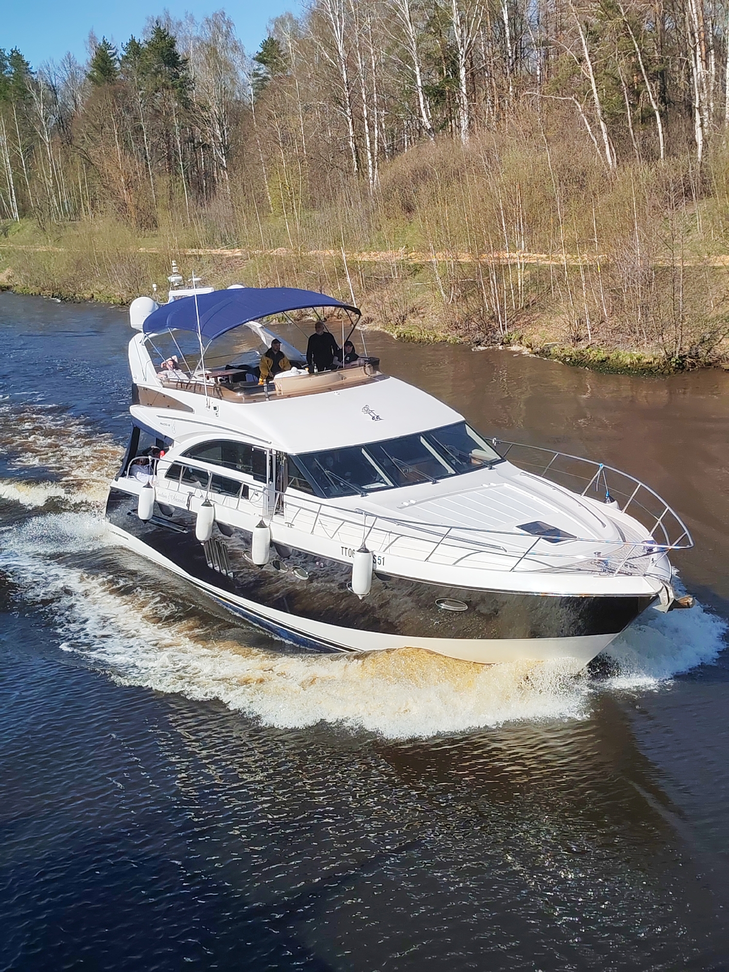 A white and dark blue yacht is moving on a river in the Орево district of Moscow, Russia. The yacht has the letters "TT06" and "551" on the front, along with "Southern & Alexander" printed on the side. Two individuals are present on the upper deck under a blue canopy. The surrounding environment includes a dense forest lining the riverbanks under a clear sky.