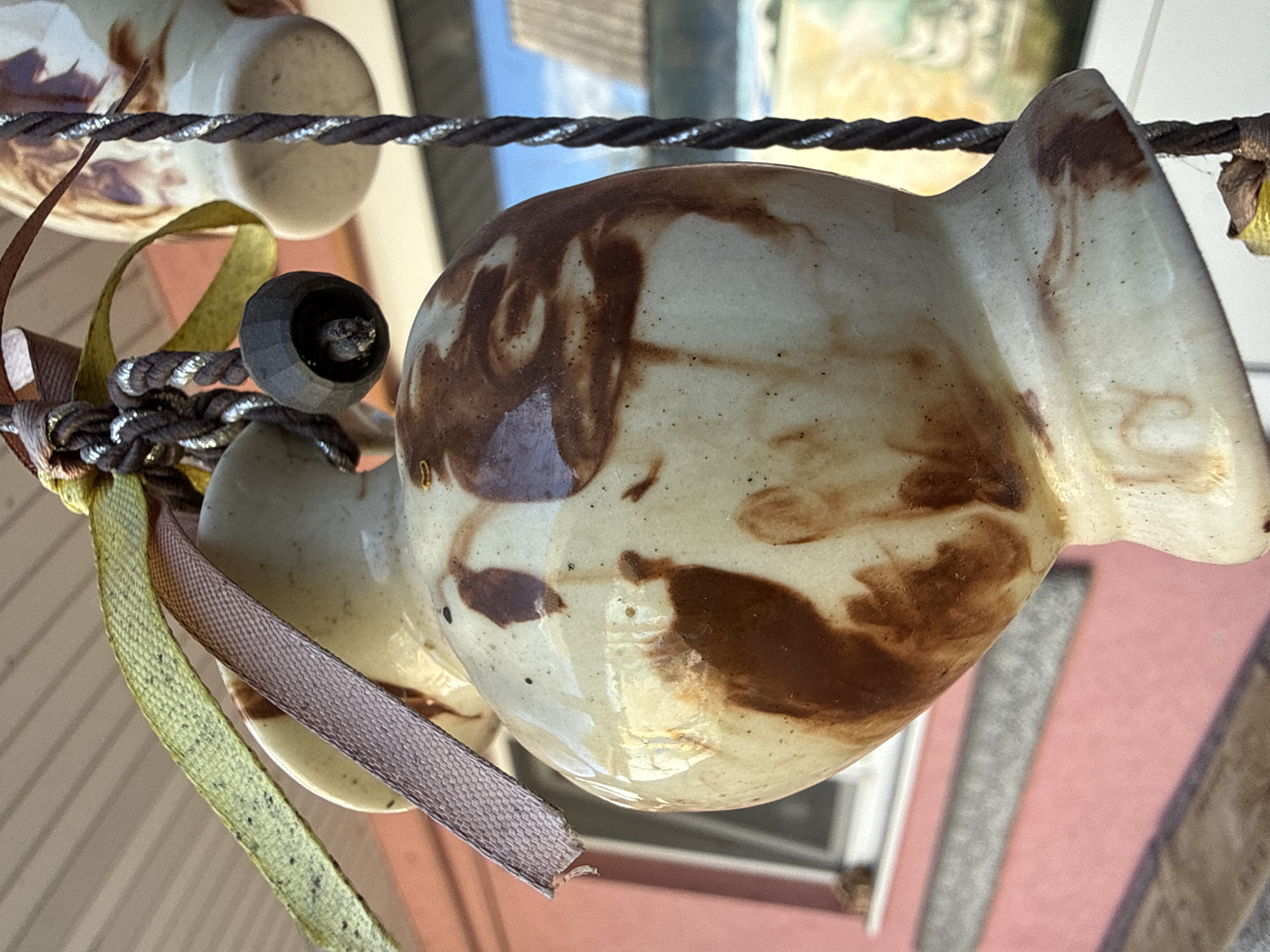 An outdoor, eye-level, close-up shot features a ceramic hanging ornament. The ornament is cream-colored with brown marbling and appears to be made of clay. It is decorated with two thin ribbons, one pink and one green, tied around its neck. A twisted brown cord with silver threads is attached to the top of the ornament. The background shows a pink building with a window, a gray ledge, and a sliver of the sky. The lighting suggests it is daytime. The image was taken in Okhtyrka, Ukraine.