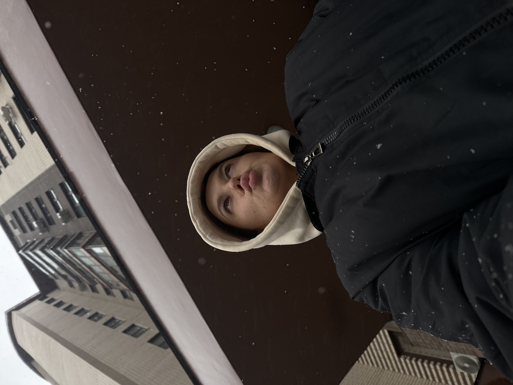 A low-angle, rotated image captures a young person from below, looking upwards and to the left with a pouting expression. The subject is wearing a light-colored hooded sweatshirt with the hood up, underneath a black winter puffer jacket with a prominent metallic zipper. Small white snowflakes are visible on the jacket and floating in the dark, reflective background, indicating current or recent snowfall.
The background primarily consists of a dark, glossy surface, likely a window or a wet, reflective street, which mirrors sections of tall, modern buildings. These buildings, reflected on the left side of the frame, feature multiple stories, numerous windows, and appear to be constructed from light-colored materials, suggesting an urban or suburban setting. The overall scene conveys a cold, winter day. There is no visible text. The location context provided is Akulovo (Gp Kubinka), Russia.