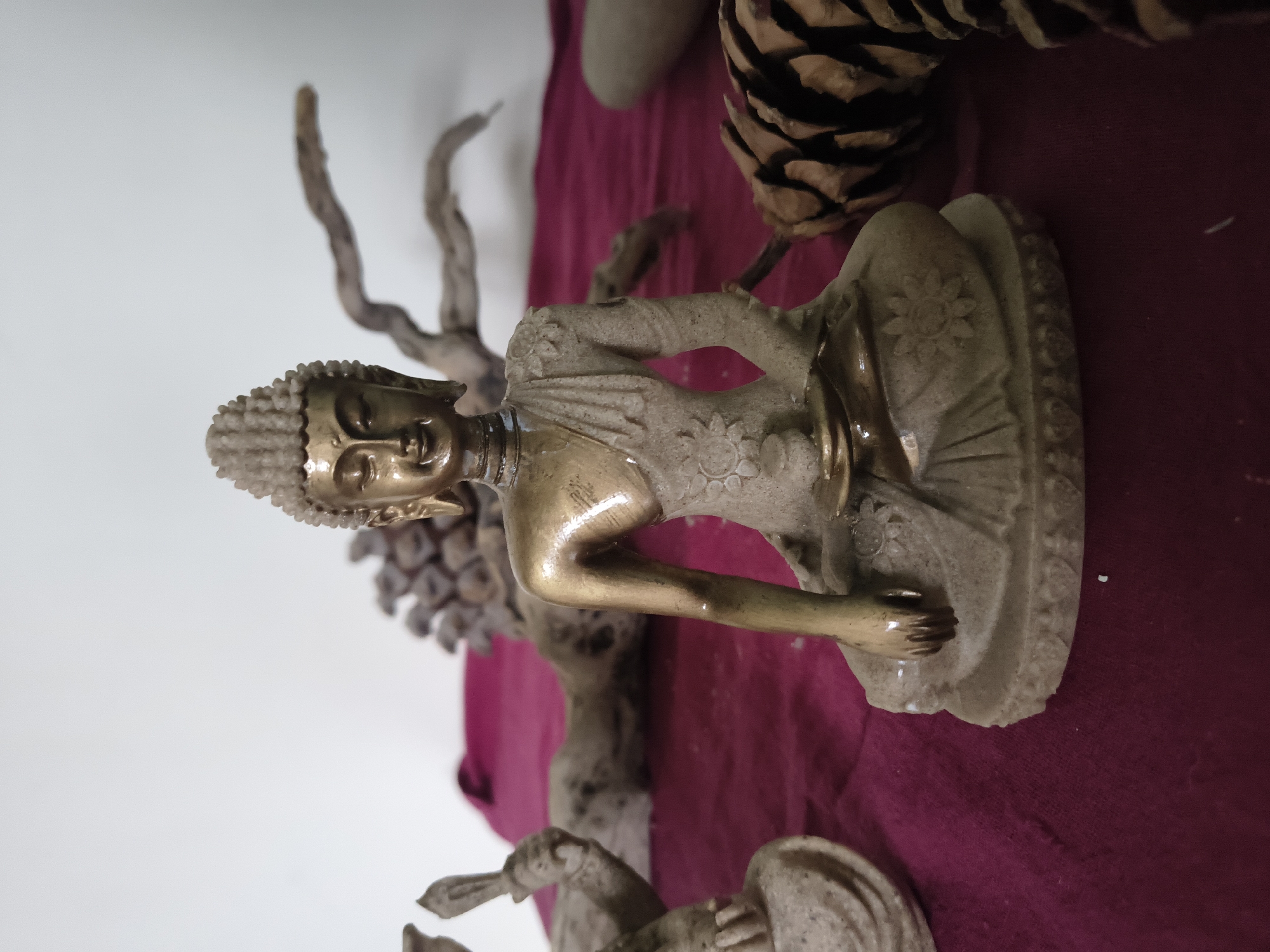 A close-up, eye-level, outdoor shot features a small, seated Buddha statue as the primary subject, positioned centrally on a deep crimson fabric. The statue is crafted from a light-colored material, possibly stone or resin, with a metallic, possibly bronze or brass, finish on the upper torso and head. Intricate floral patterns adorn the statue's robe. The Buddha is depicted in a meditative pose, with eyes closed and a serene expression.
In the background, out of focus, are elements that suggest a naturalistic or decorative setting. To the left, another statue fragment, possibly part of a similar Buddha figure, is visible. Behind the main statue, a piece of driftwood, with gnarled branches, is arranged horizontally. Further back, a cluster of what appears to be pine cones or similar natural objects is present. The backdrop is a plain white wall, contributing to a clean, minimalist aesthetic. The lighting is soft, creating subtle highlights and shadows on the Buddha statue. The image does not depict any people or provide any specific geographical markers that would indicate a location in Haini, India.