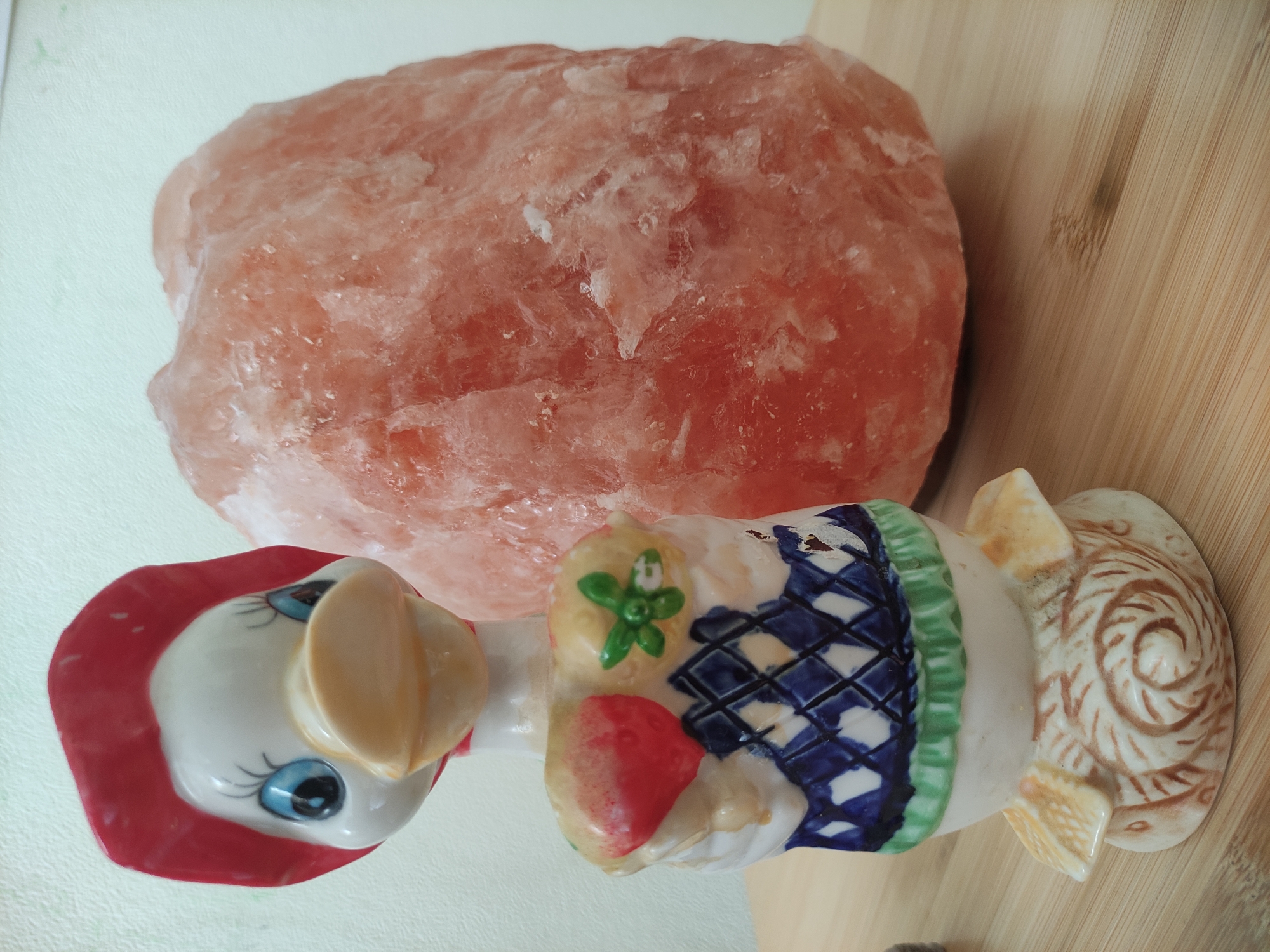 An overhead shot captures a still life arrangement featuring a large, crystalline, pinkish-orange rock and a ceramic figurine. The rock, resembling a Himalayan salt chunk or lamp, is irregularly shaped with a textured, almost translucent surface, showing variations in its coral and peach hues. It occupies the upper and central-right portion of the frame.
To the lower left of the rock is a detailed ceramic figurine. The figurine depicts a stylized character, possibly a bird or duck, with a white body, prominent blue eyes outlined with black lashes, and a light beige beak. It wears a broad, matte red hat that frames its face. The body of the figurine is adorned with a dress-like pattern: a band of dark blue and white diamond crosshatching, a green ruffled trim, and a small red, heart-shaped or strawberry-like detail with a green flower-like pattern above it. The base of the figurine is bulbous and decorated with intricate brown, swirling patterns on a lighter background, resting partially on a light wooden surface.
The background consists of two distinct surfaces: a smooth, light green or off-white surface to the left, and a wooden surface with visible grain lines to the right, which also extends under the figurine's base. No activity is taking place, and there is no visible text. The lighting is bright and even, suggesting an indoor setting.