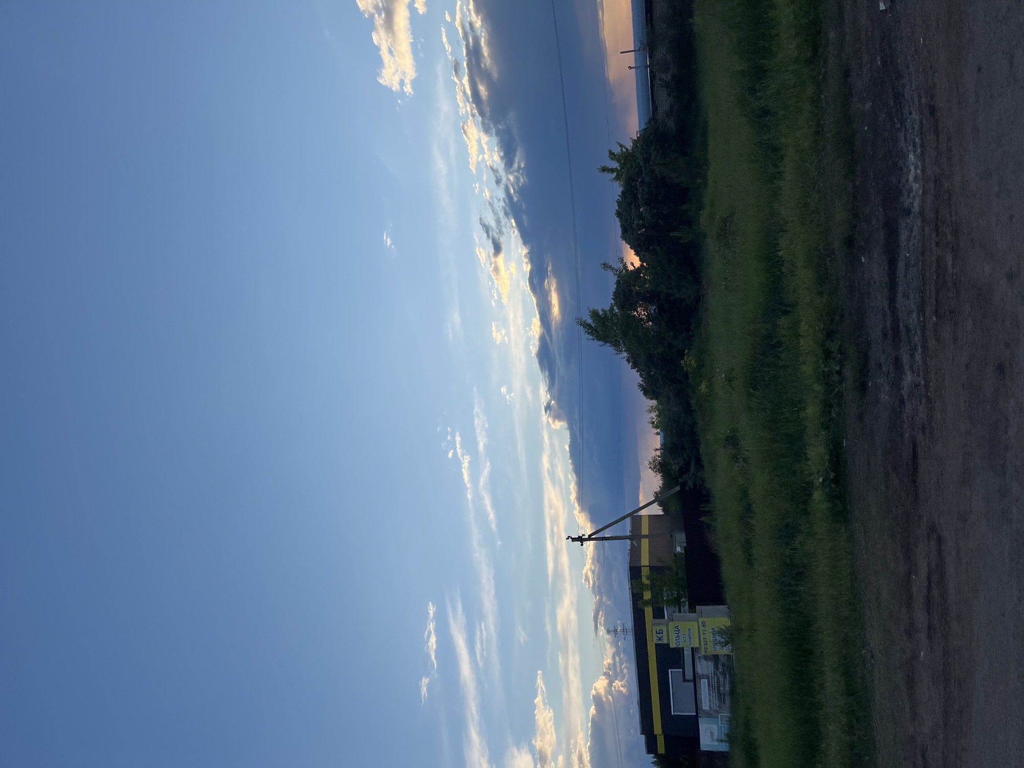 The image depicts a scene in Chaikovsky, Russia. The view is oriented toward a blue sky with wispy, elongated clouds tinged with sunlight along their edges. Along the right side of the image is a narrow strip of green vegetation, appearing to be a grassy area or field. A dirt or gravel road runs along the bottom right edge. Adjacent to the vegetation is a low-rise building with a black and yellow facade, marked with signage in Russian. A utility pole with power lines is visible near the building. The light suggests it is either dawn or dusk.