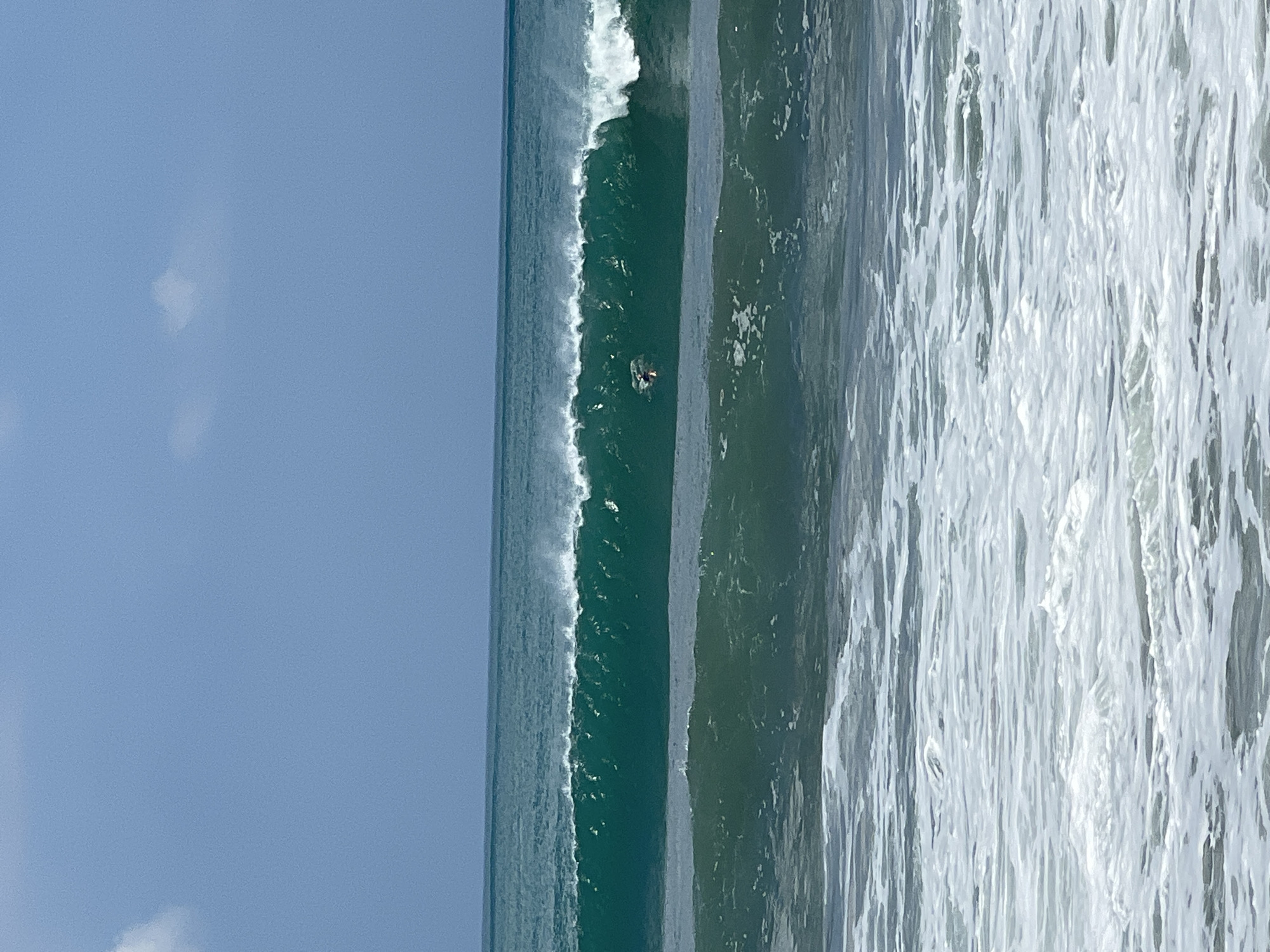 A surfer rides a breaking wave in the ocean off the coast of Kuta, Indonesia. The wave is a vibrant emerald green, with white foam cresting as it barrels. The sky above is a clear, pale blue with a few wispy clouds. In the foreground, the ocean is calmer, with gentler waves and foam washing ashore. The image captures a moment of action and the natural beauty of the ocean.