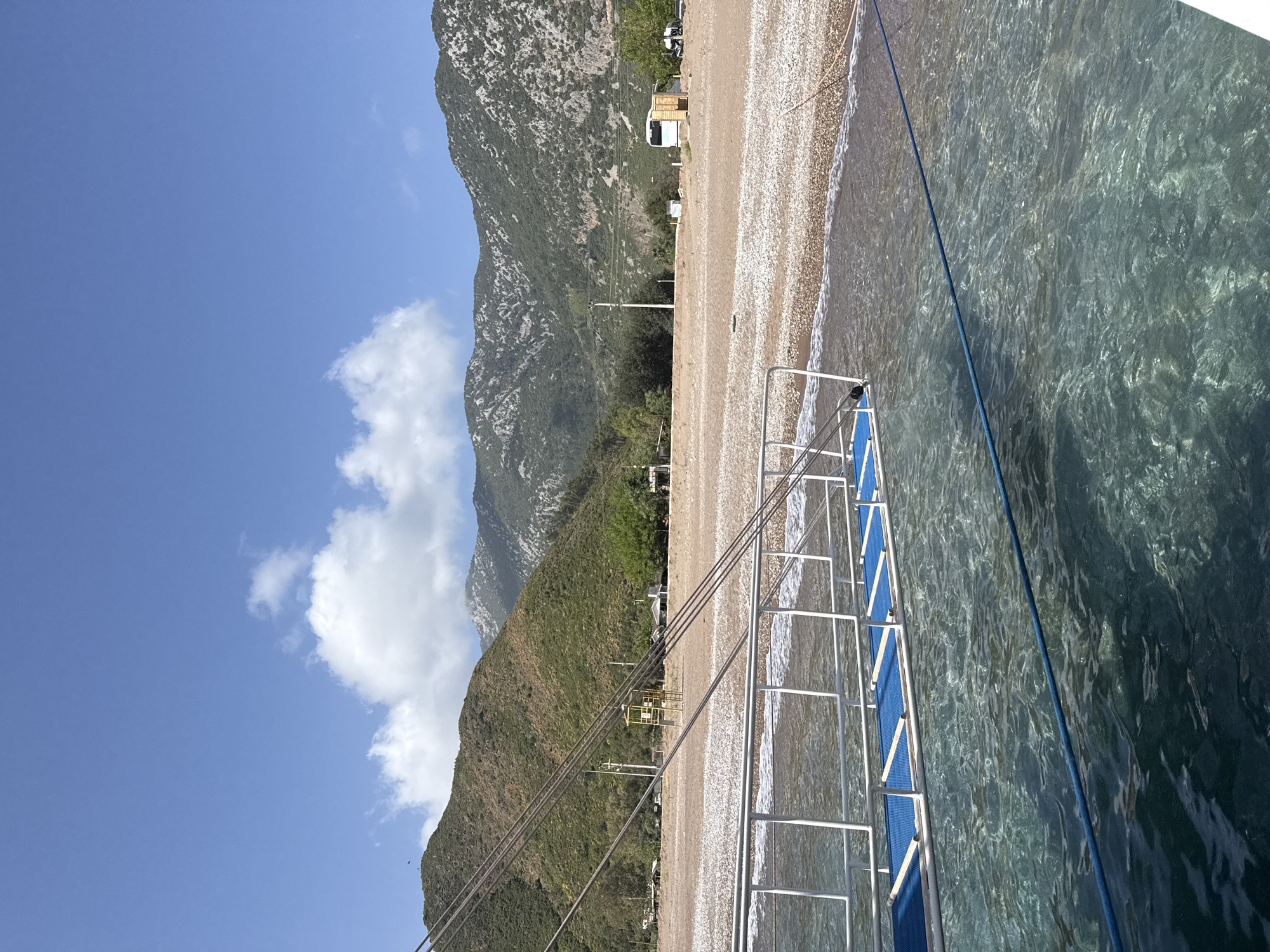 The image captures a coastal scene in Kumluca, Turkey, viewed from the deck of a boat. The foreground features the clear turquoise waters of the Mediterranean Sea with a boat ladder extending into the water. Adjacent to the sea is a pebble beach with small waves gently lapping at the shore. Beyond the beach, the terrain rises sharply into forested mountains that dominate the landscape. A few buildings and vehicles are visible along the coastal strip. The sky is blue with scattered white clouds, suggesting a clear day. Power lines also run up the mountain side.