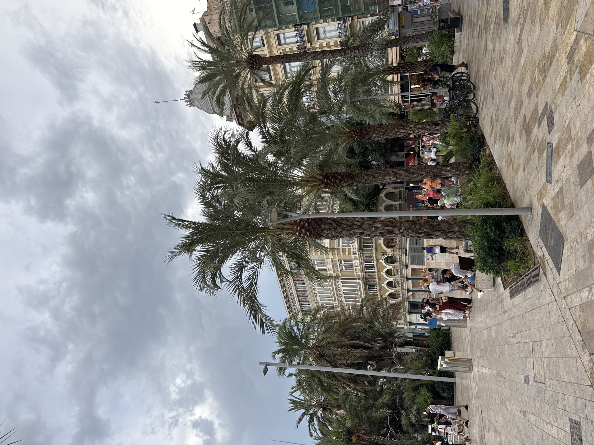 The image shows a street scene in Valencia, Spain, with a cloudy sky overhead. Palm trees line a pedestrian walkway paved with light-colored tiles. In the background, several ornate buildings with multiple stories and balconies stand tall. Some of these buildings appear to be under construction or renovation, with scaffolding visible on one. A group of people are scattered throughout the scene, some walking, others standing and conversing. One man is seated on a bench, looking at his phone. Bicycles are parked near a patch of greenery. The overall atmosphere suggests a public square or promenade within a city.