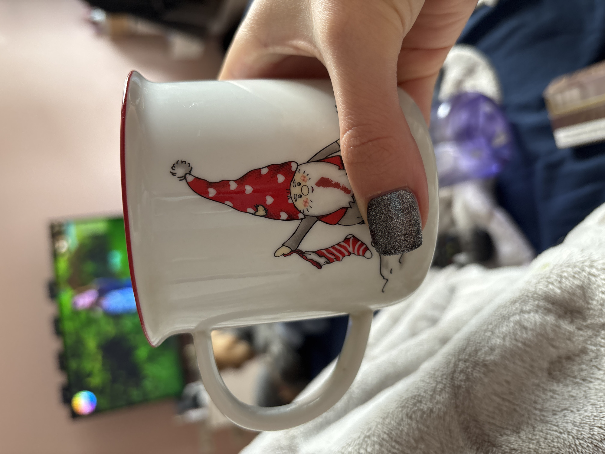 A hand is holding a white mug with a red rim and a cartoon illustration of a gnome-like figure wearing a red and white polka-dotted hat and holding a striped stocking. The background includes a television with a colorful screen and a white fluffy blanket. The location is not discernible from the image.