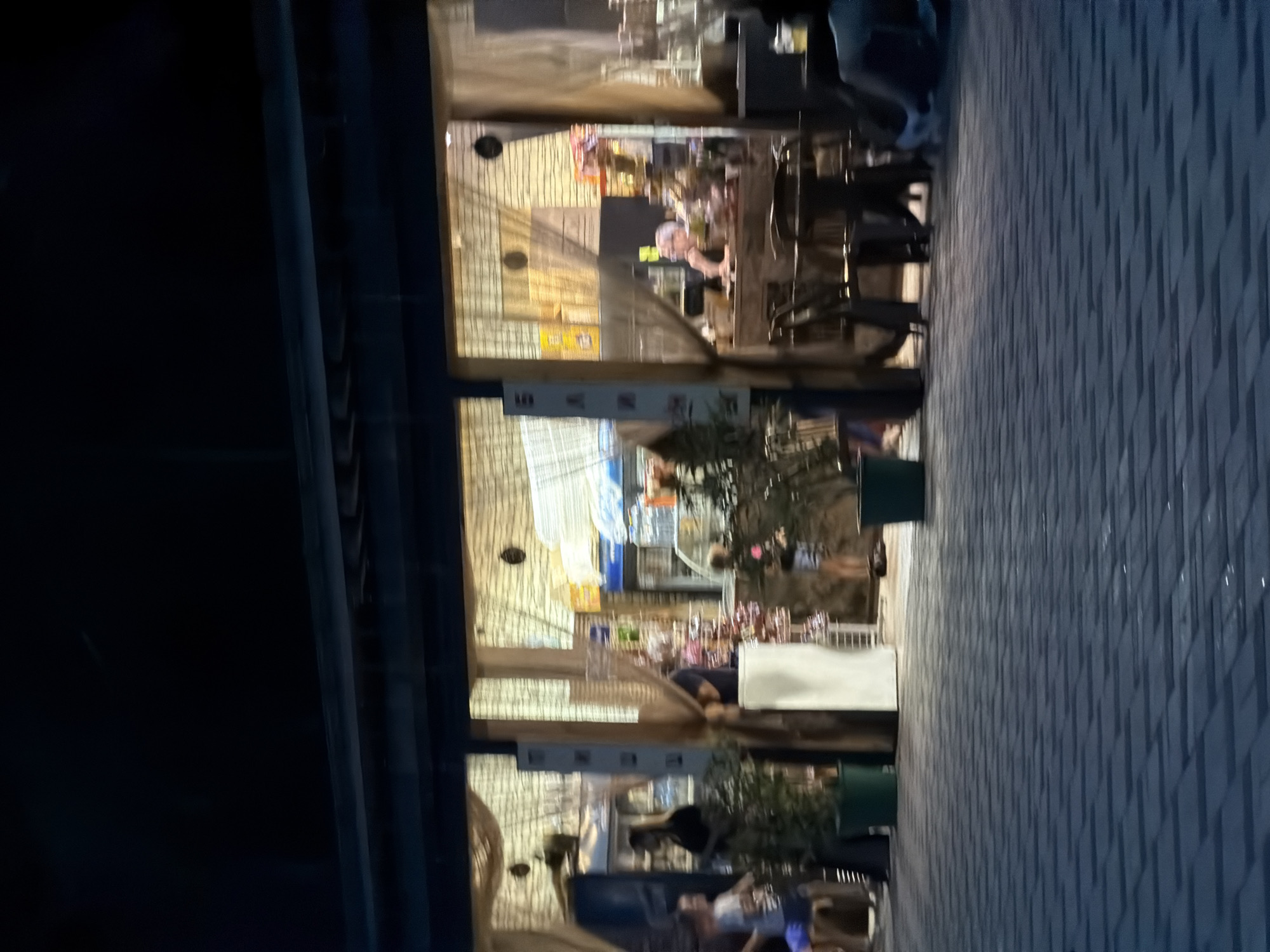 The image depicts a nighttime view of an outdoor seating area in front of several small shops or kiosks in Partenit, Ukraine. The foreground is paved with rectangular stone tiles, laid in a patterned arrangement. In the midground, there are several covered structures with wide openings, revealing interiors illuminated by artificial light. These interiors appear to house retail spaces, with shelving, products, and refrigerators visible. Two individuals, a woman and a man, are visible inside one of the kiosks, engaged in what appears to be service or transaction. Another person, possibly a customer, is seen interacting with the man. In the far right of the midground, a person is seated at a table outdoors, with a beverage in front of them. The background is dark, suggesting it is nighttime and the sky is not visible. The overall impression is a lively, open-air commercial area, likely a promenade or a marketplace, at dusk or after dark.