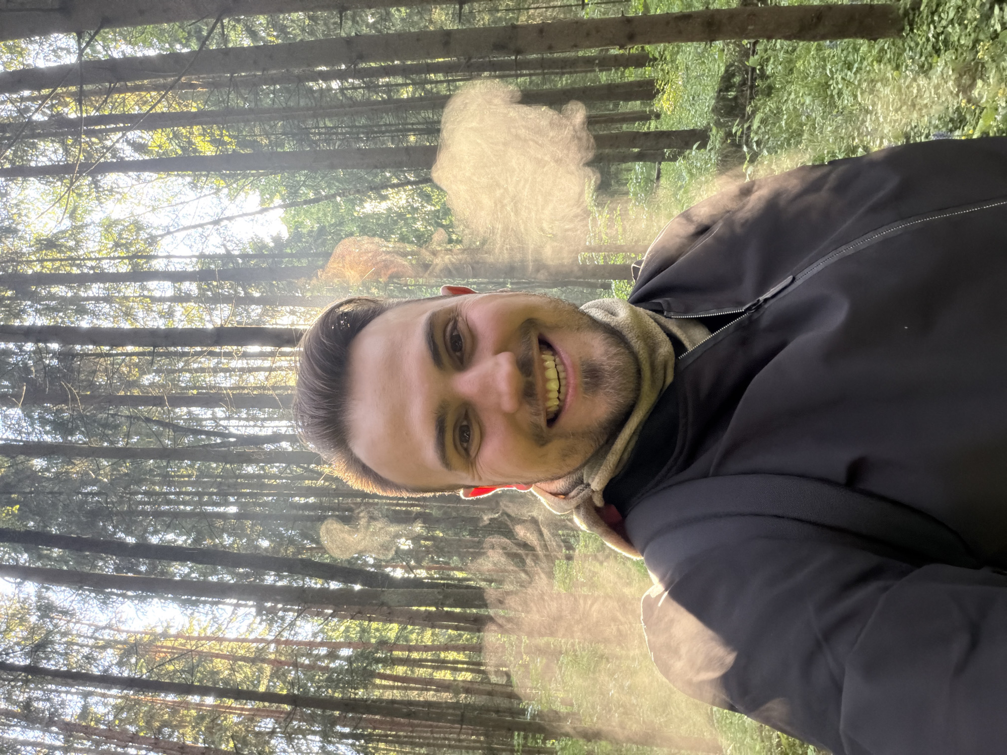 A man with short brown hair and a trimmed beard is smiling at the camera in a selfie taken in a forest. He is wearing a dark-colored jacket and a light-colored scarf or turtleneck. Light is streaming through the trees, creating a hazy or misty effect in the air. The trees are tall and slender, and the forest floor is covered in green vegetation. The location appears to be Troitsk, Russia, judging by similar landscapes found in the area.