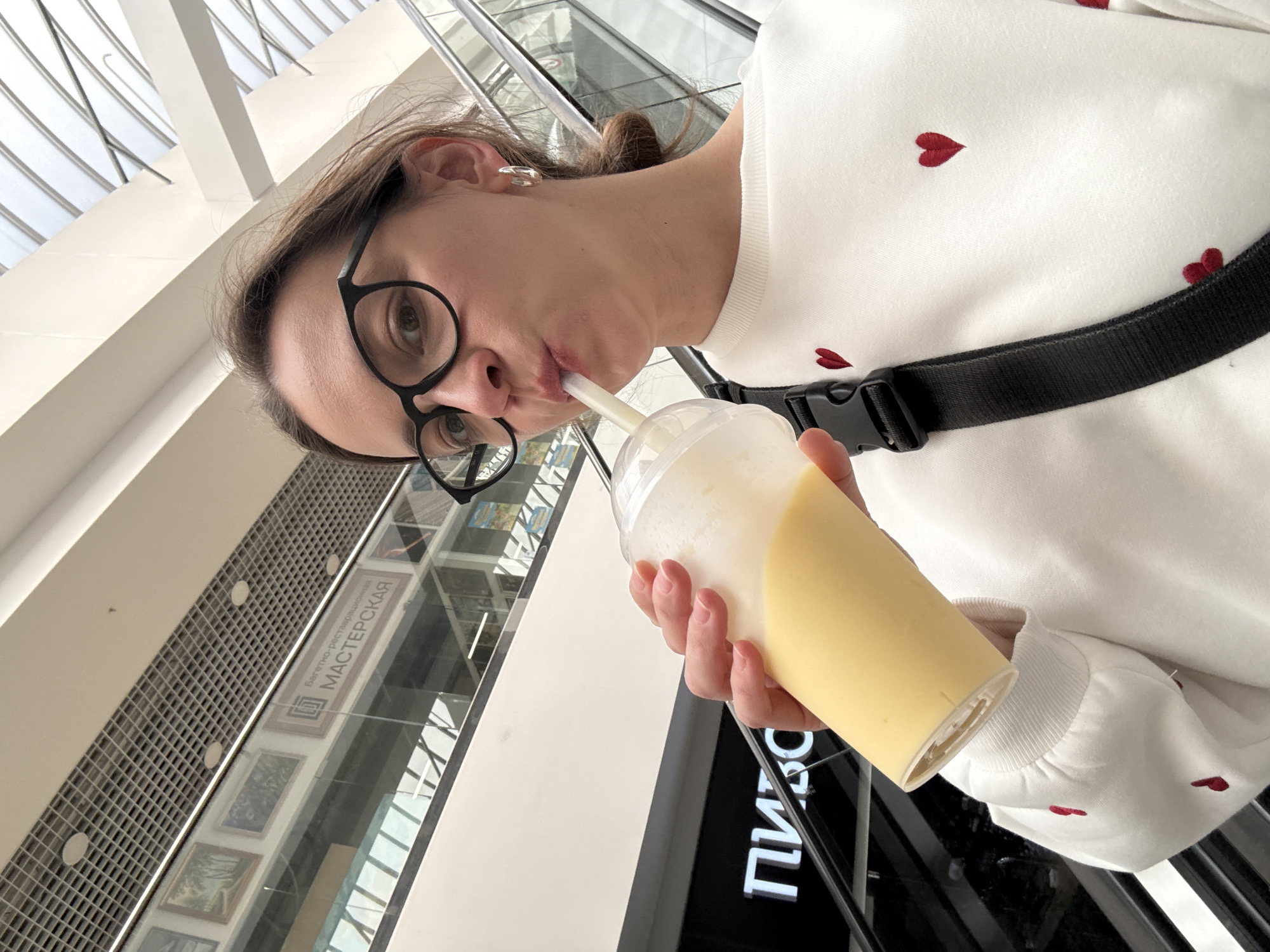 A woman with glasses is drinking from a plastic cup with a straw. She is wearing a white sweater with small red hearts. She has a black strap across her chest. The location appears to be a shopping mall in Izhevsk, Russia. The words "МАСТЕРСКАЯ" and "ПИШ" are visible in the background.