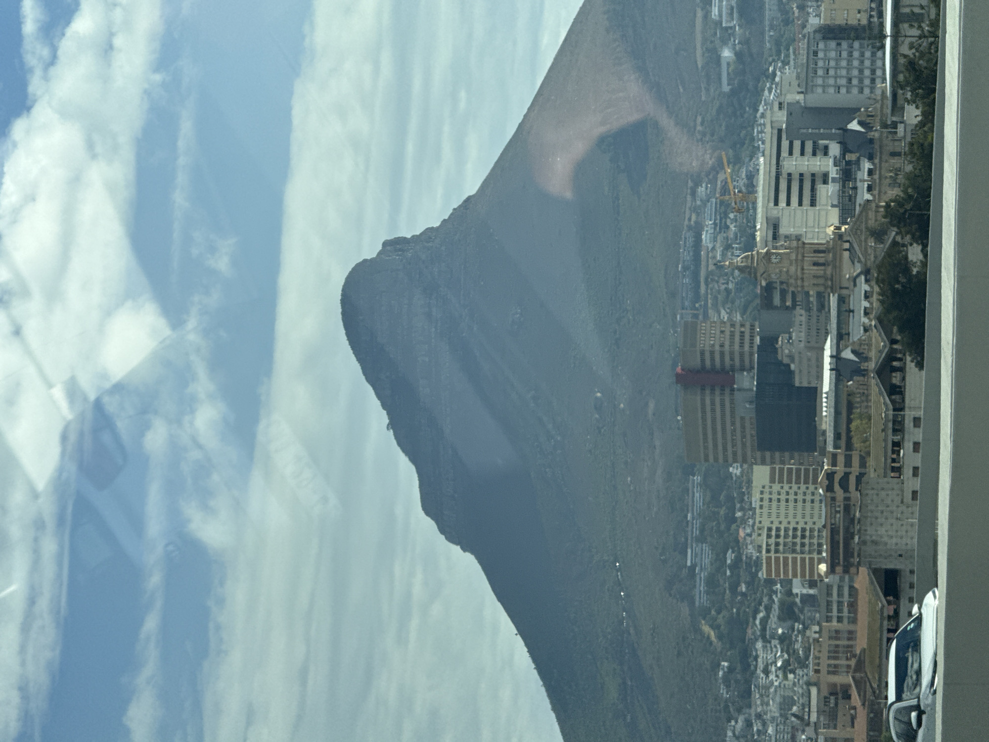 The image depicts a view of Table Mountain in Cape Town, South Africa, seen through a window. The sky has scattered white clouds. Table Mountain itself is dark green with a distinctive flat top. The city of Cape Town is visible at the base of the mountain, with a mix of buildings, including high-rise structures, with visible vegetation. A white car is partially visible on the right edge of the image.