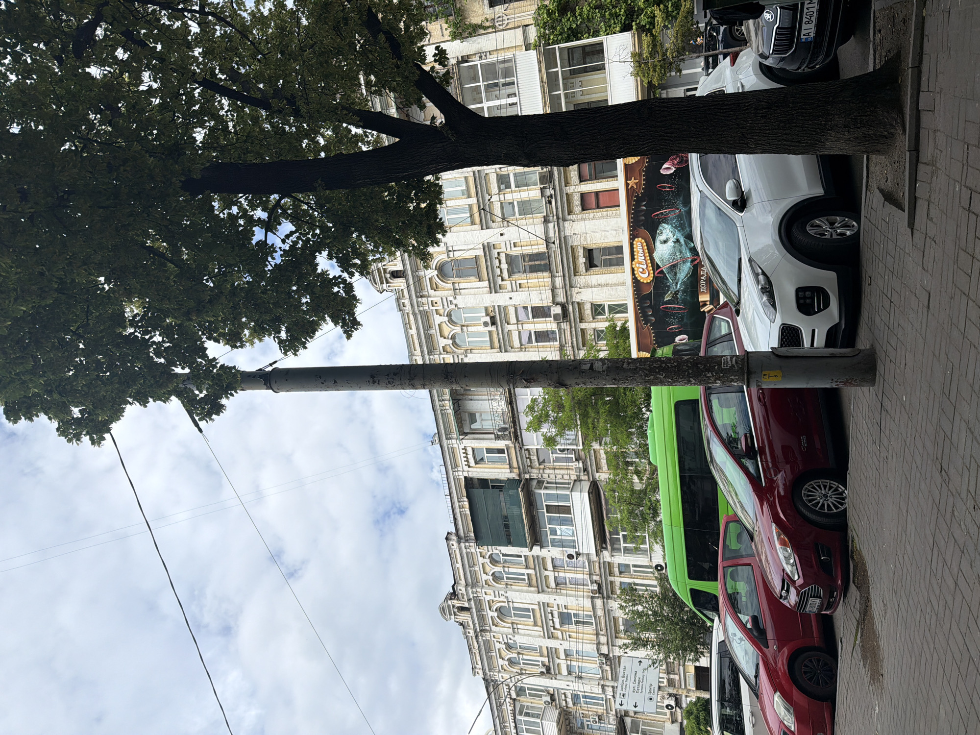 The image captures a street scene in Kyiv, Ukraine, likely in a historic district, given the architecture of the building. In the foreground, several cars are parked along a sidewalk made of brick pavers. The cars include a white Kia Sportage, a dark-colored Skoda, and two red cars, a hatchback and a sedan. Behind the cars, a bright green bus is visible. The building behind the bus is a multi-story structure with ornate architectural details, featuring multiple windows and balconies, some with visible air conditioning units. A large tree with lush green foliage partially obscures the building and the sky. The sky is overcast with scattered clouds. A utility pole stands prominently in the center of the frame, with wires stretching across the sky. On the building facade, there is a large banner with an advertisement that features a fish jumping through hoops. Signage is visible on the left, with text in Ukrainian, indicating directions to "Заліз. Вокзал" (Railway Station) and "Центр" (Center), suggesting this is a public area with transportation access. The overall impression is a typical urban street in Kyiv, characterized by older architecture, parked vehicles, and street infrastructure.