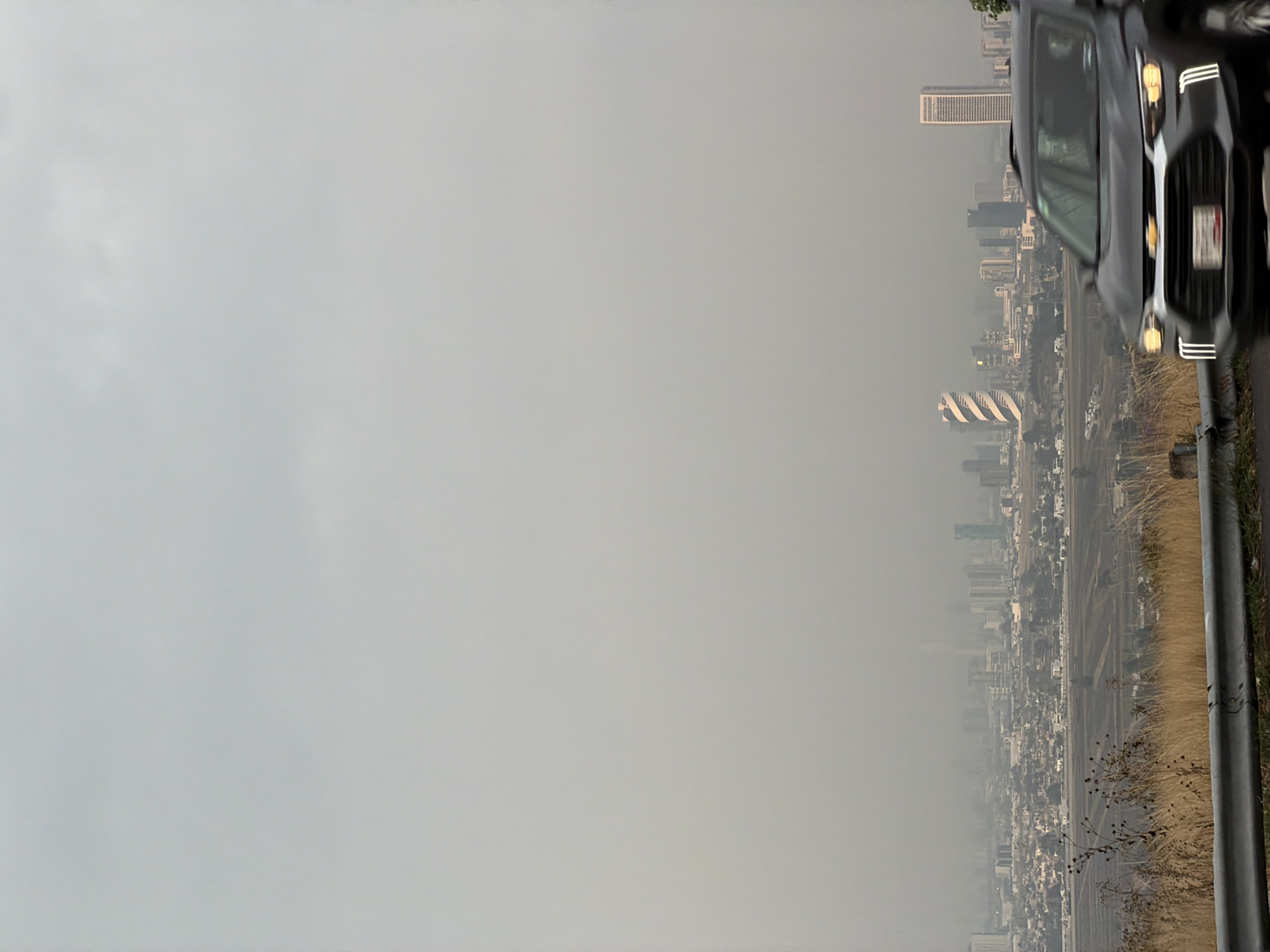 The image shows a cityscape obscured by smog, possibly taken from a high vantage point. A portion of a dark car is visible on the right edge of the frame. The city appears to be Santa Clara Ocoyucan, Mexico, based on the presence of distinctive buildings like the spiral-shaped Arcos Bosques 1 tower and other recognizable structures. A roadside barrier is also visible. The sky is overcast and hazy.