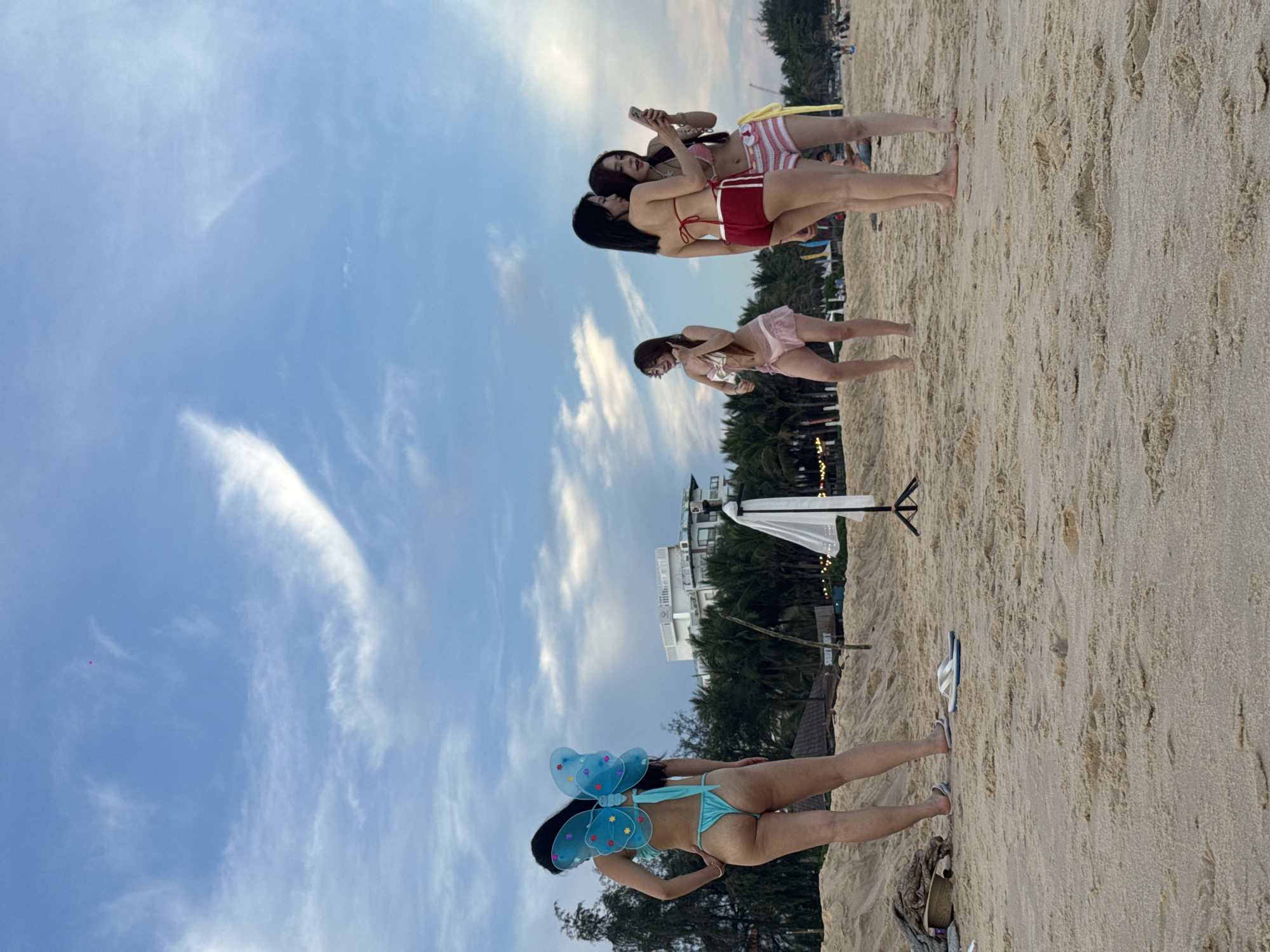 On a sandy beach in Phu Thuy Ward, Vietnam, four young women are gathered near the water's edge. The sky above is a soft blue, with wispy white clouds. In the foreground, one woman stands with her back to the camera, wearing a turquoise bikini and blue butterfly wings. To her right, three other women are grouped together. Two of them are looking at a smartphone, one holding it, while the third stands slightly apart, holding a drink. They are all dressed in bikinis and shorts. In the background, palm trees line the shore, and a multi-story building with balconies is visible. The beach appears to be a public area with a few scattered items, including a tripod with a white cloth, a straw hat, and a pair of flip-flops.