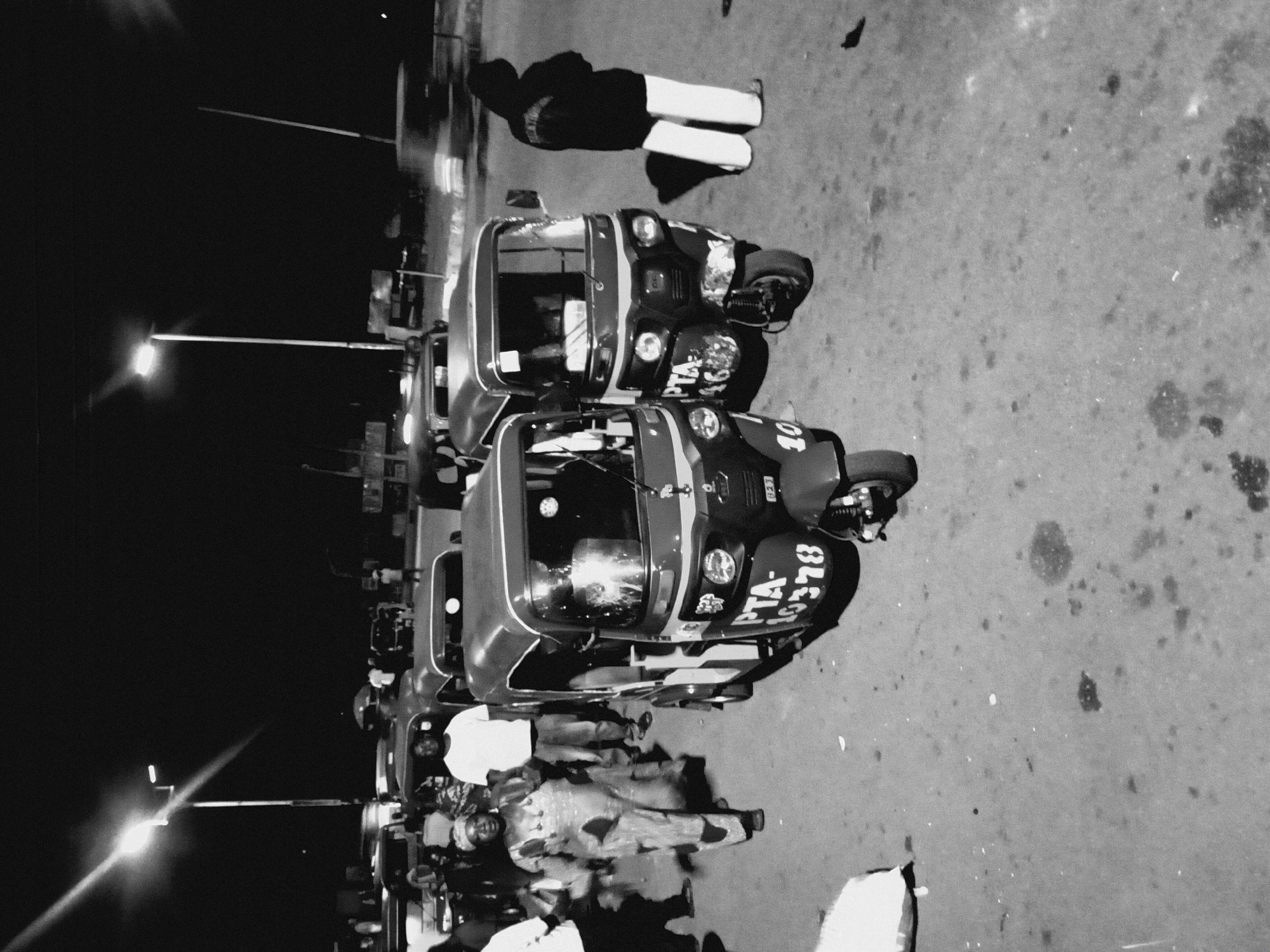 The black and white image depicts a street scene at night in Bukuru, Nigeria. A row of three auto-rickshaws, locally known as "Keke Napep," are parked along the roadside. Each vehicle has a visible license plate with the prefix "PTA-". Pedestrians and drivers stand around the vehicles. Overhead streetlights cast a glow on the scene and the road. The asphalt road surface shows uneven patches and dark stains. One person standing near the auto-rickshaws appears to be wearing white trousers. The environment suggests a commercial or gathering area.