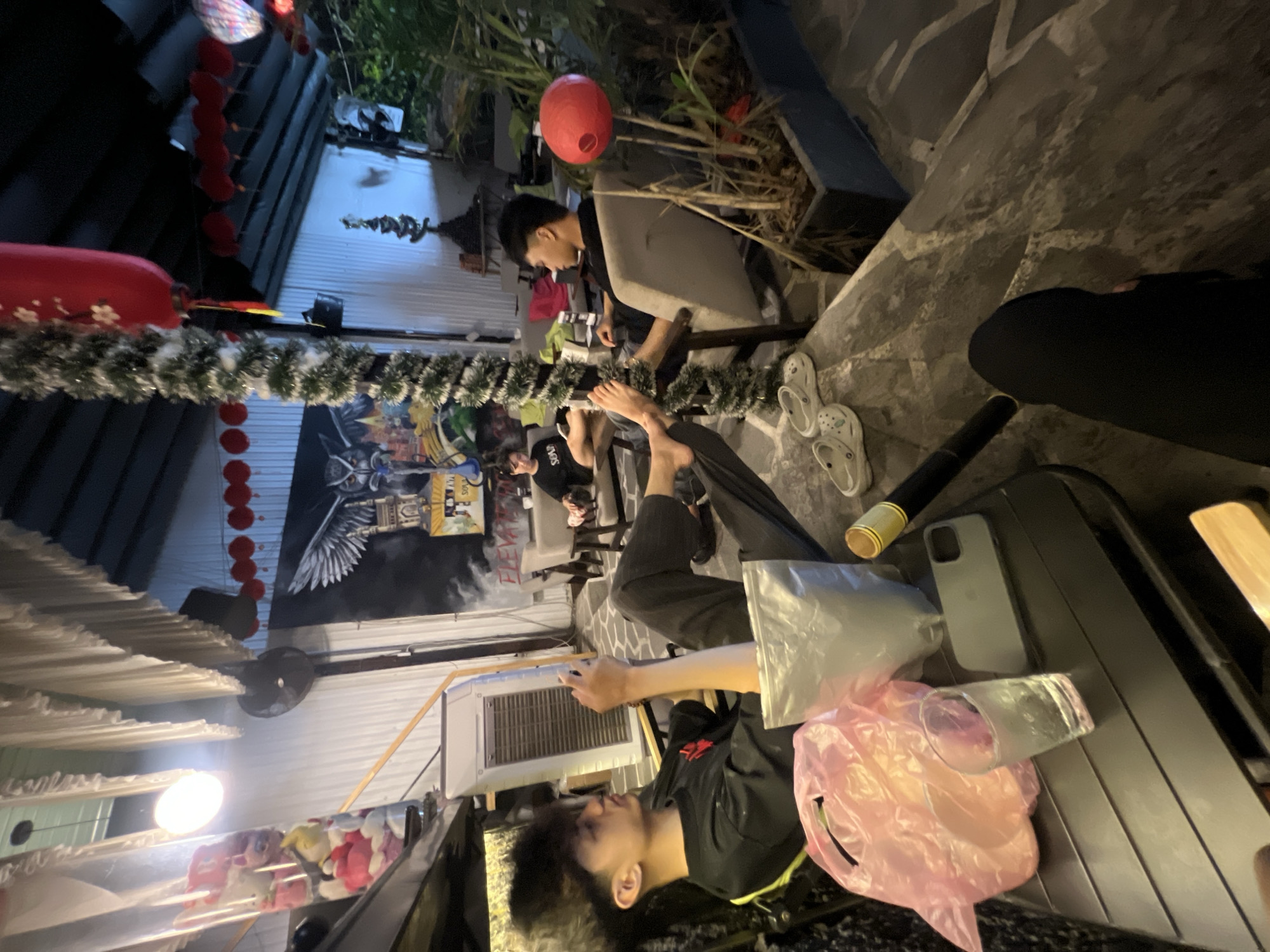 The image depicts an outdoor scene in Đà Nẵng, Vietnam, possibly a courtyard or patio. Several people are present, including at least two men seated and another reclining. One man is wearing dark pants and a shirt. A black suitcase sits close to him. Footwear is visible near a seating area. Decorations, including red lanterns, adorn the space. The light is dim, and the image has a slightly rotated perspective. A table nearby contains a water bottle, a mobile phone, and several plastic bags. An air conditioning unit is visible on the building. A mural decorates the building's exterior.