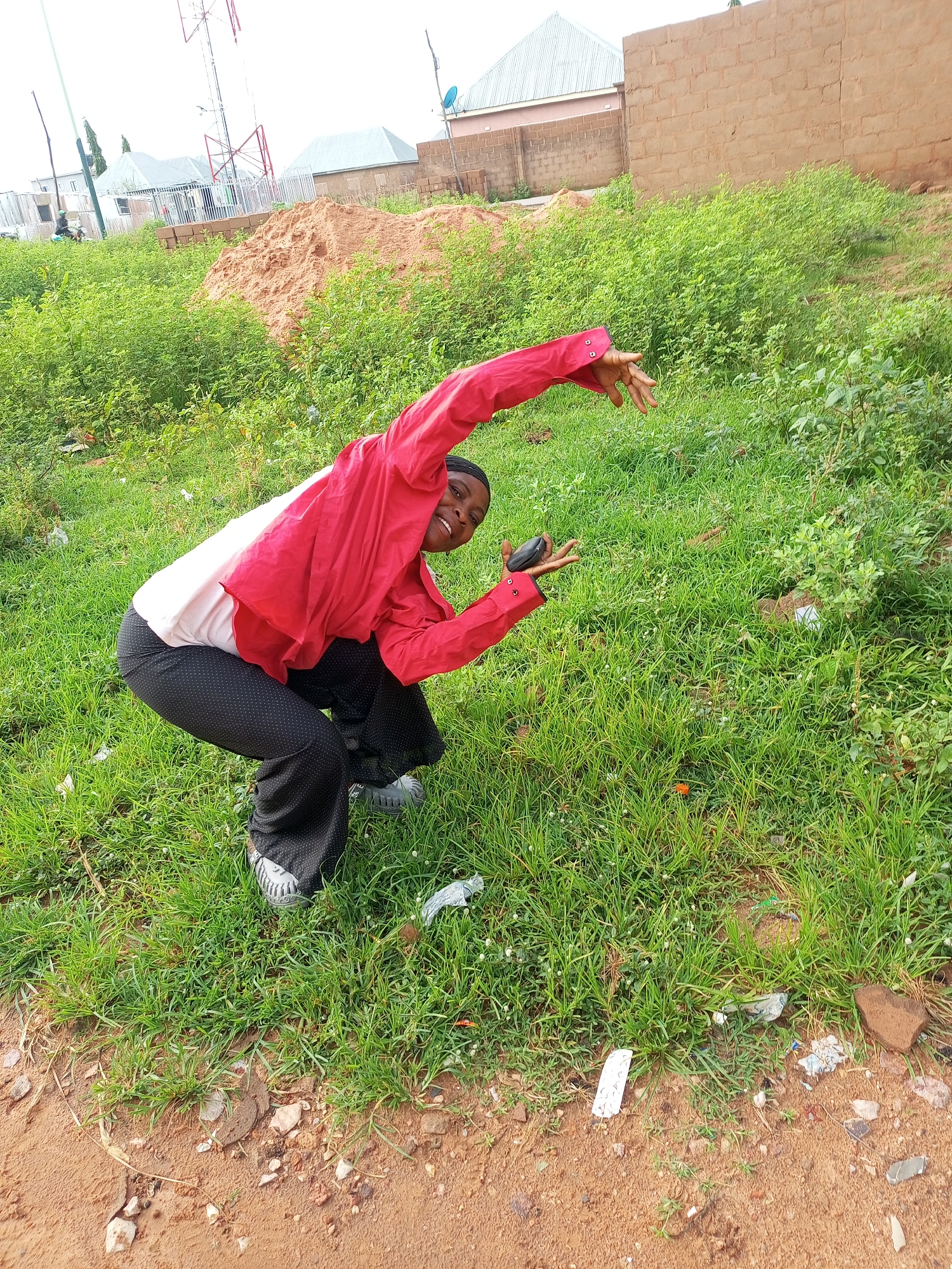 A young Black woman is crouching in a grassy, unpaved area. She is wearing a bright red, collared shirt with the sleeves rolled up and a white t-shirt underneath. Her pants are black with a white polka dot pattern. She is looking towards the camera with a wide smile and has a black object, possibly a phone, in her left hand. Her right arm is extended outwards with her hand open. In the background, there are some buildings, a telecommunications tower, and a pile of orange sand. The ground is covered in green grass with some scattered litter. The sky is overcast and bright. The location is identified as Tempure, Nigeria.