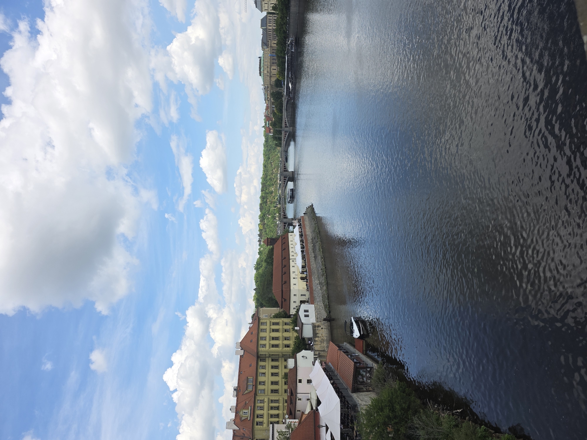 The image shows a cityscape along a river, likely Prague, Czech Republic. Buildings with red tile roofs and light-colored facades line the riverbank. Bridges span the river in the distance, and the sky is filled with clouds. The river reflects the sky and surrounding buildings.