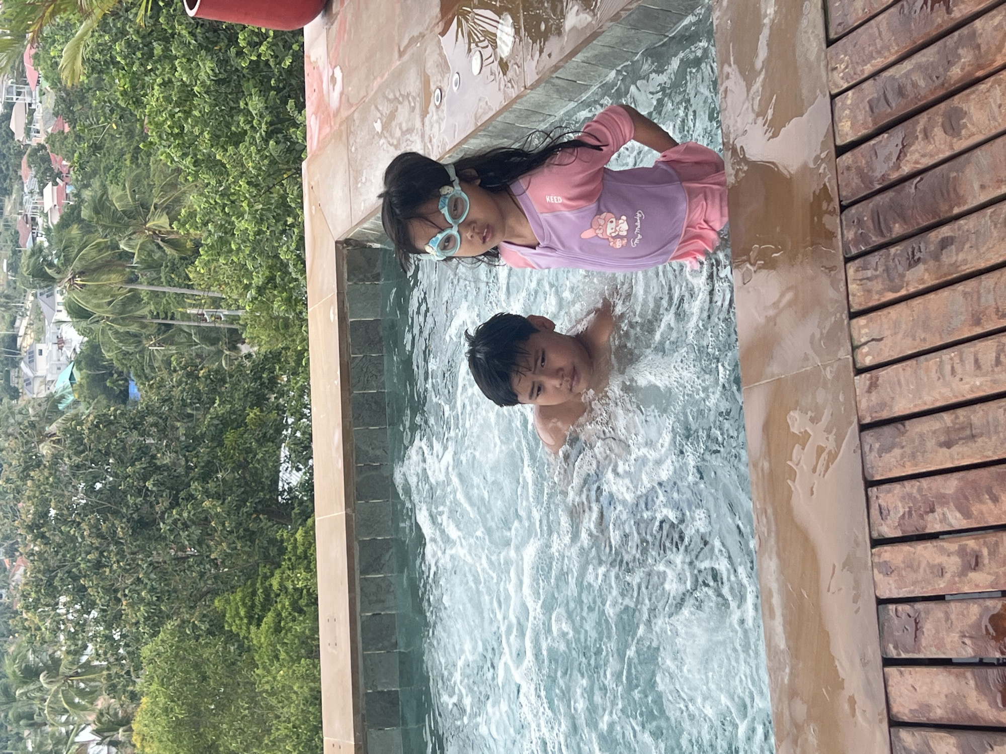 Two children are enjoying a hot tub overlooking a tropical landscape in Ko Samui District, Thailand. The boy, on the left, is partially submerged in the bubbling water, looking towards the right. The girl, on the right, stands facing the viewer, wearing blue swimming goggles and a pink and purple swimsuit with a cartoon character on it. The hot tub is made of tiled walls and has a wooden deck surrounding it. In the background, lush green trees and houses are visible on a hillside, suggesting a scenic, elevated location. The weather appears overcast.