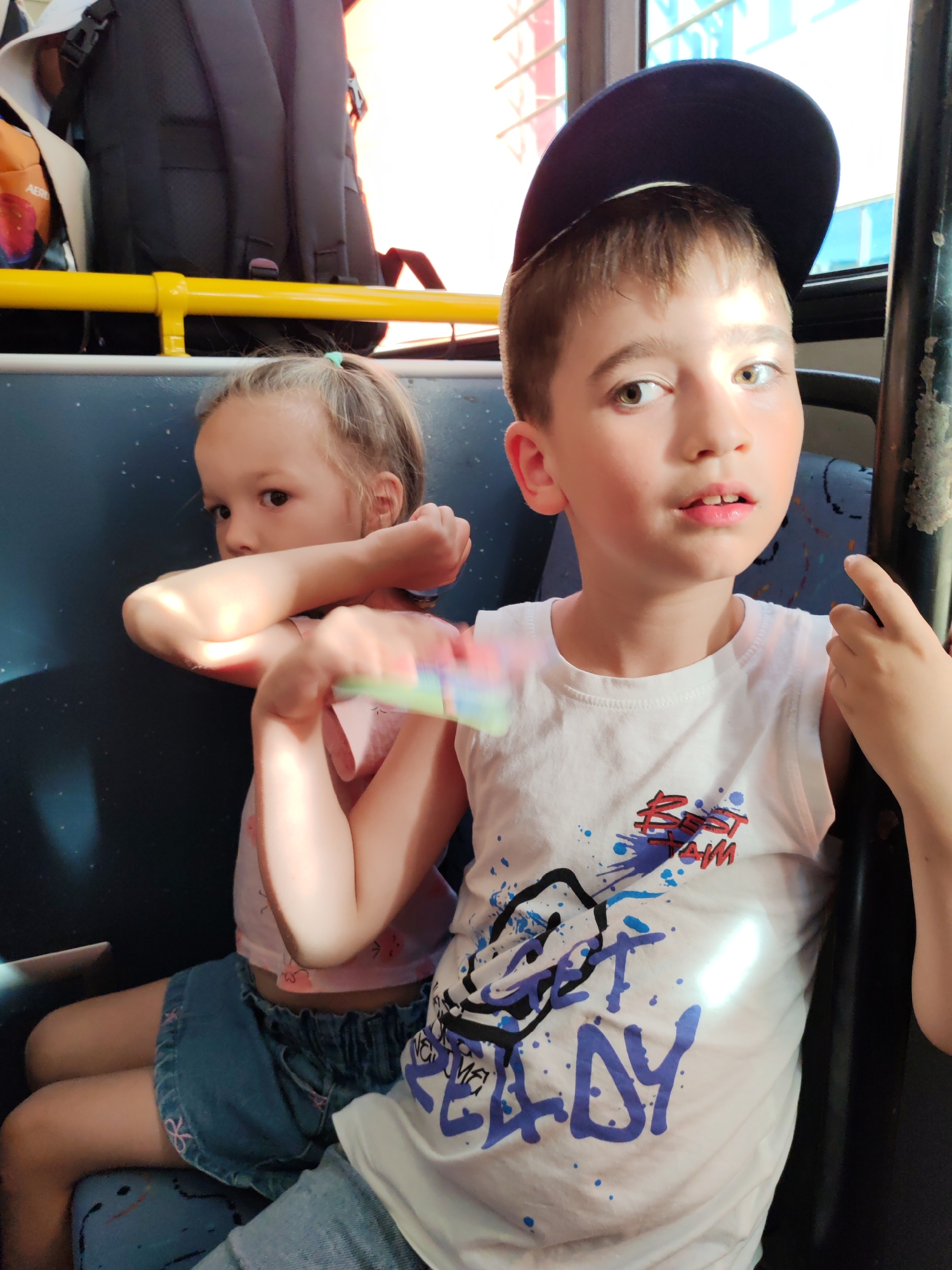 The image shows two children, a boy and a girl, sitting on a bus. The boy is in the foreground, wearing a white sleeveless shirt with a graphic design that includes the words "BEST TEAM" and "GET READY". He has short brown hair and is wearing a dark baseball cap. He is looking towards the camera with a surprised expression. The girl is sitting next to him, slightly behind and to the left. She has blonde hair tied in a ponytail and is wearing a pink top and a denim skirt. She is looking away from the camera with a neutral expression. She is holding a colorful object, possibly a toy, in her hands. The setting appears to be the interior of a bus, with blue seats and a yellow handrail visible. Sunlight is streaming through the windows, casting shadows. The scene suggests a casual bus ride. The location is identified as Khrabrovo, Russia.