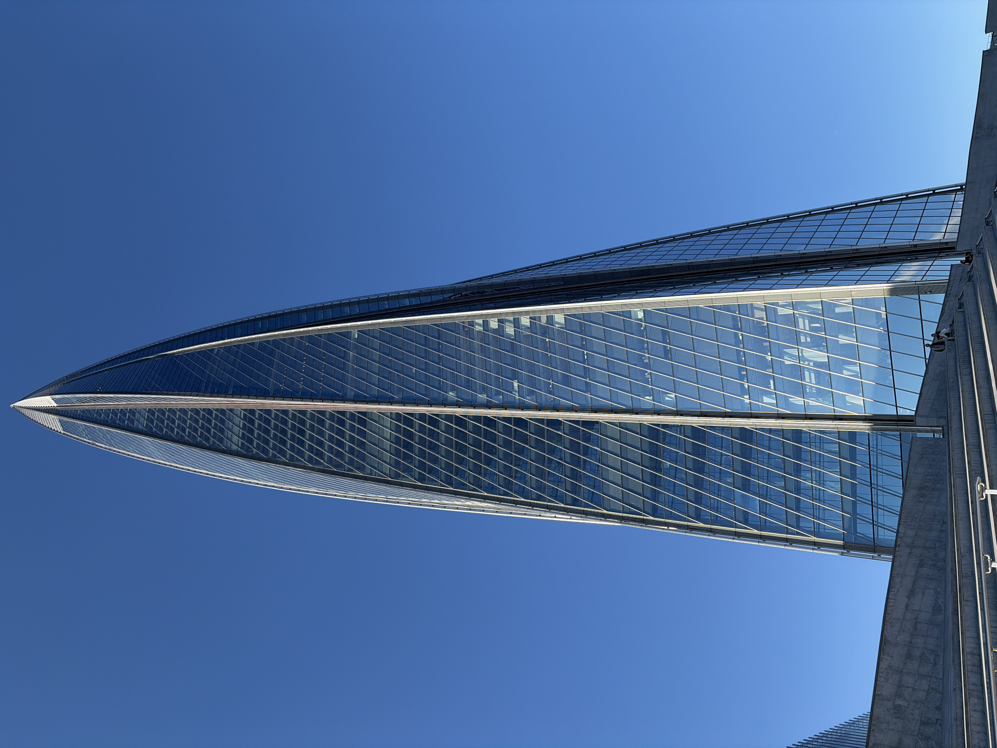 The image shows a low-angle, full-shot view of the Lakhta Center, a skyscraper in Saint Petersburg, Russia, against a clear blue sky. The building's unique, spiraling architectural design is prominent, with its glass facade reflecting the sky and creating a dynamic visual effect. In the foreground, there are concrete steps and tiered platforms, with a few individuals visible sitting on the steps. The scene is bathed in sunlight, highlighting the modern architecture and vastness of the sky.