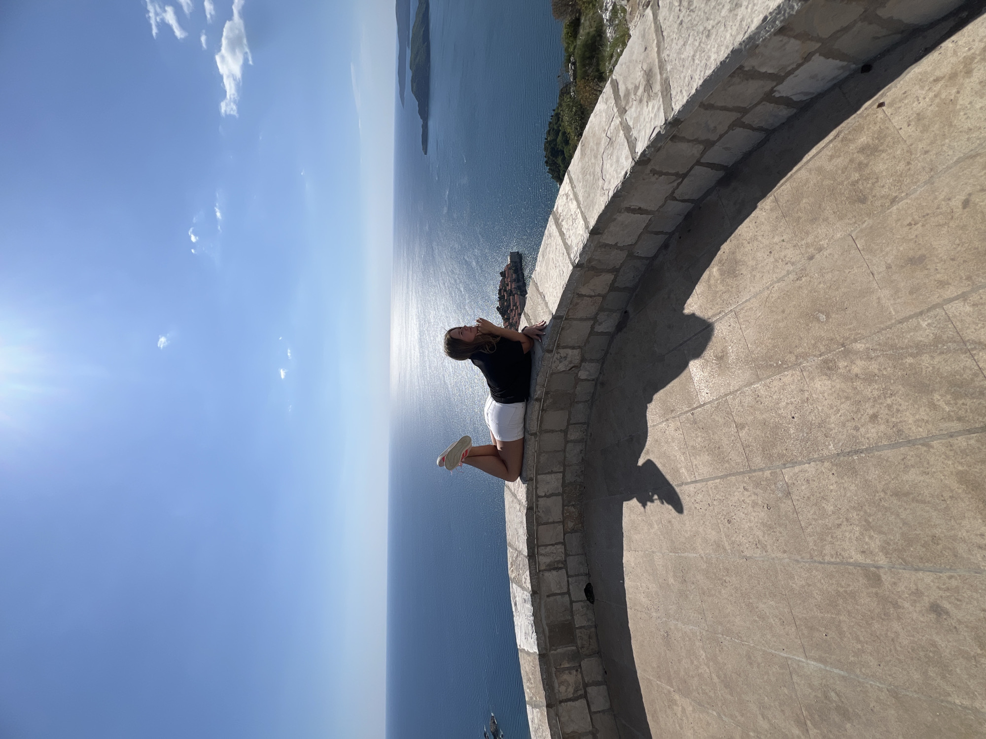 The image depicts a young woman lying on the edge of a stone wall, overlooking the Adriatic Sea. She is wearing a black shirt and white shorts with red shoes. The wall appears to be part of a circular structure, possibly a fortification or observation point. The location is suggested to be in Sveti Stefan, Montenegro, judging by the coastal scenery. The sky is blue with scattered clouds.