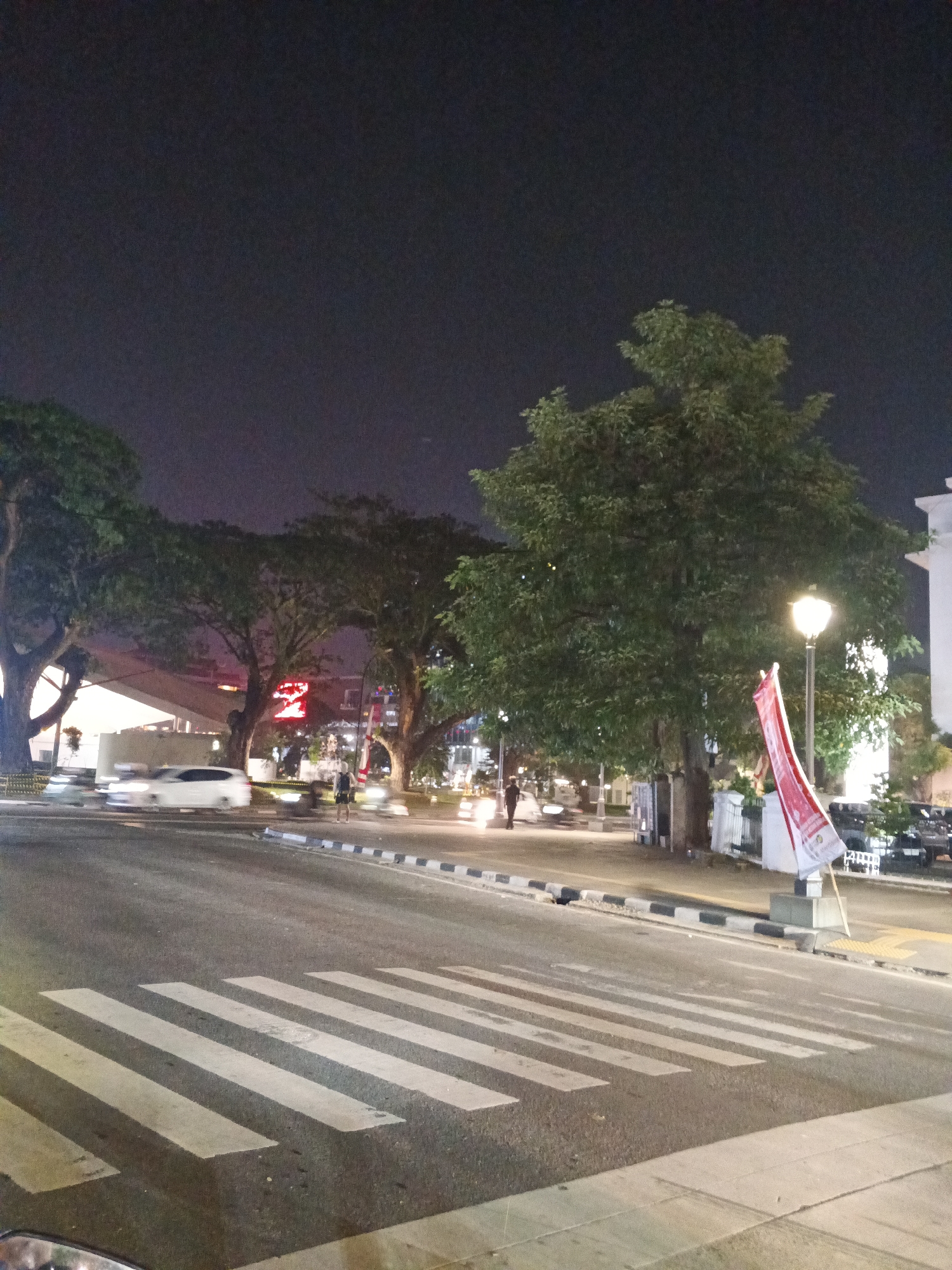 The image captures a nighttime scene on a city street in Medan, Indonesia, featuring a pedestrian crossing in the foreground. The sky is dark, indicating it is likely late evening or night. The street is paved, and the pedestrian crossing is marked with white stripes. To the right of the crossing, a street lamp illuminates the area, casting a warm glow. A red and white flag, possibly the Indonesian flag, is mounted on a pole near the lamp post, slightly unfurled.
Further down the street, trees with lush green foliage line the sidewalks, their branches reaching upwards. The trees appear mature and well-established. Behind the trees, there are indications of buildings and structures, some with illuminated signage, suggesting a commercial or public area. A prominent red neon sign with the number "2" is visible between two large trees.
Several vehicles are present on the road. A white car is seen moving from left to right in the middle ground, captured with motion blur. Other vehicles, including what appear to be parked cars, are visible on the right side of the street. A lone pedestrian is standing on the sidewalk to the left of the street lamp, facing away from the camera. The overall environment suggests a well-lit urban setting, possibly a busy intersection or a parkway within the city. The presence of the flag and the style of the trees are consistent with locations in Indonesia.