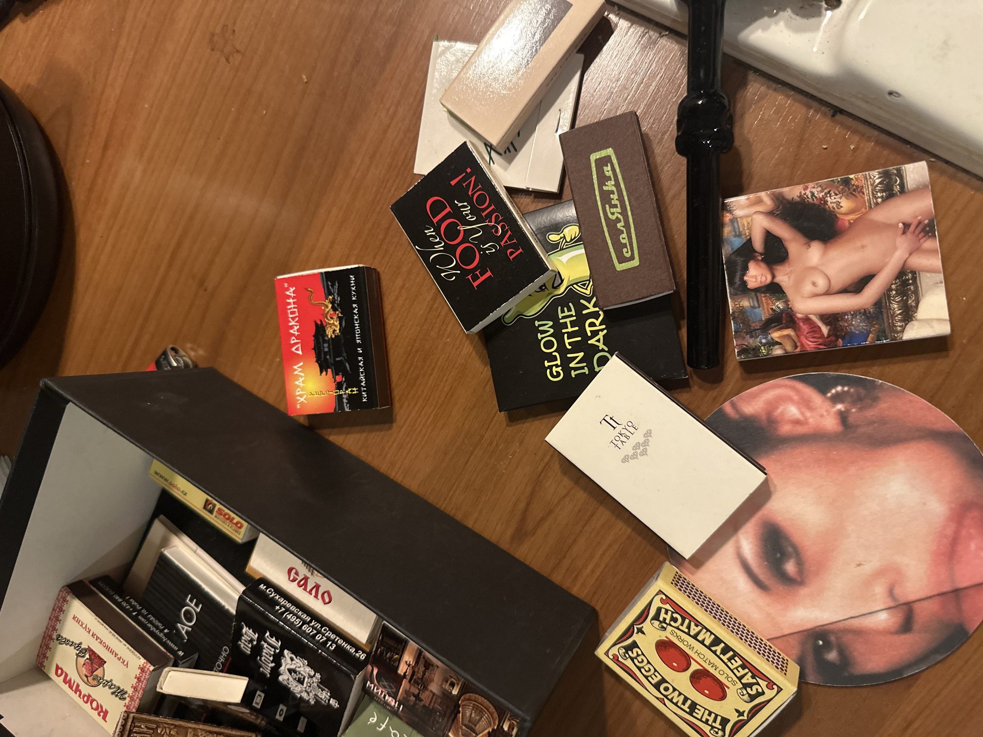 The image shows a wooden table with various items scattered on it. In the upper left corner, there is a black box filled with matchboxes. Some of the matchboxes have text in Russian, including "КОРЧМА" and "КИТАЙСКАЯ И ЯПОНСКАЯ КУХНИ". There are also matchboxes with images of food and people. In the center of the image, there is a matchbox with the text "When Food is Your Passion!". To the right of this, there is a matchbox with the text "GLOW IN THE DARK". Below this, there is a matchbox with the text "солянка". In the lower left corner, there is a matchbox with the text "THE TWO EGGS SAFETY MATCH". There is also a circular item with a woman's face printed on it. To the right of this, there is a small book or pamphlet. In the lower right corner, there is a matchbox with an image of a nude woman. A black object, possibly a tool or utensil, lies across the center of the table. The lighting suggests an indoor environment, possibly a home or a restaurant. The scene does not provide specific information to determine its location in Moscow, Russia, beyond the presence of Russian text on some items.