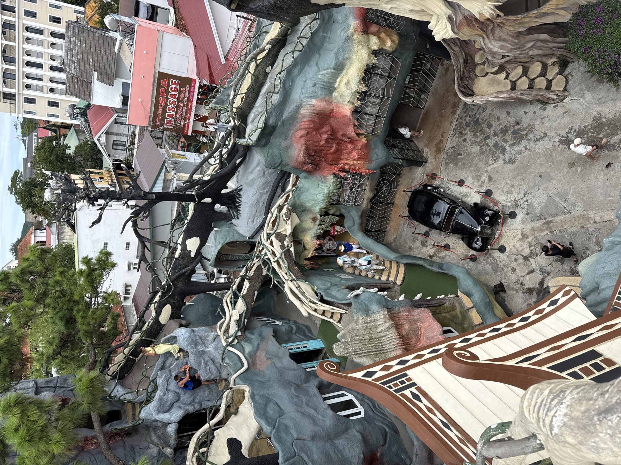 A high-angle view captures a bizarre architectural scene in Da Lat, Vietnam. The architecture appears whimsical and somewhat grotesque, resembling a cross between a Gaudi-esque structure and a treehouse. The building is built from a mixture of architectural styles and includes features such as blue-framed windows and beige patterned features.
Several people are visible in the scene. One man in a blue shirt appears to be climbing the structure. Other people are captured in groups on a lower level.
A black vintage car is parked nearby and is behind red rope stanchions. The surrounding environment consists of more ordinary buildings, some with visible signs such as a "SPA MASSAGE" advertisement. A large, bare tree trunk adds to the surreal atmosphere.