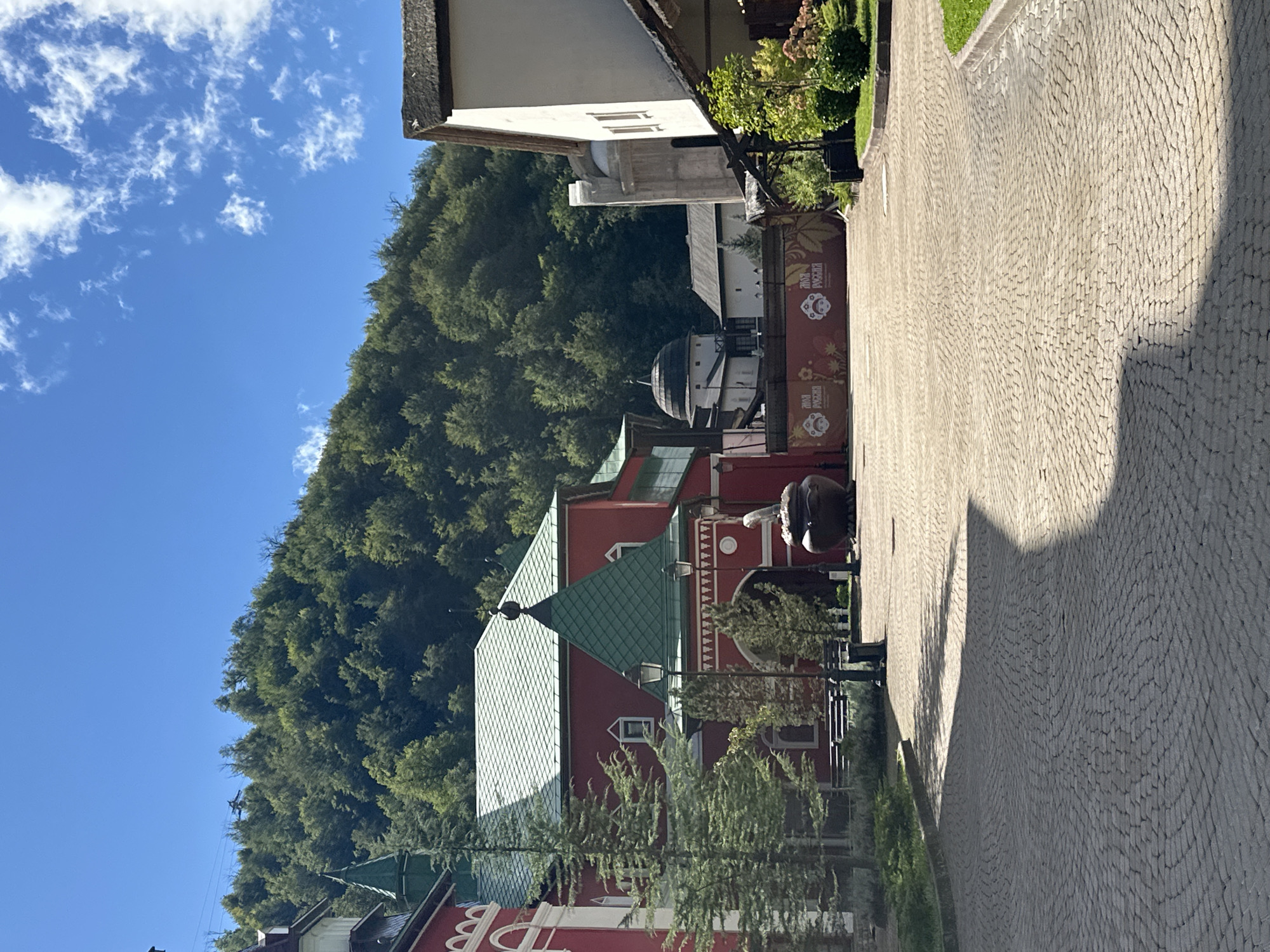 The image depicts a scene in Estosadok, Russia, on a clear, sunny day. The foreground is a cobblestone pathway with a large shadow cast across it. To the left, a red building with green accents and a pointed green roof is visible, featuring arched entrances and decorative white trim. Evergreen trees are planted near the building.
In the mid-ground, a large, dark, cauldron-like object with a white, statue-like figure on top is situated near an arched entryway. Behind the red building, other structures are visible, including a domed building and a white building with a thatched roof. A banner with Cyrillic text and imagery is displayed on a fence or barrier in the mid-ground.
The background is dominated by a lush, green, tree-covered mountainside under a bright blue sky with scattered white clouds. The overall impression is a well-maintained outdoor space, possibly a park or a cultural attraction, with traditional Russian architectural elements. There are no people visible in the image.