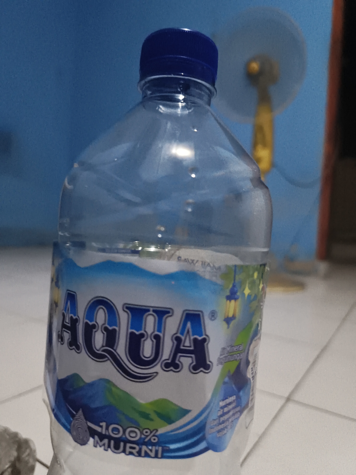 A clear plastic bottle of Aqua brand mineral water is depicted. The bottle has a blue cap and a label that features the Aqua logo, mountains, and a water droplet. The label also indicates "100% MURNI" (100% Pure). In the background, a blue wall is visible, along with a yellow fan with a light-colored oscillating head. The scene appears to be indoors, on a light-colored tiled surface. The overall lighting is dim. This image does not contain enough information to determine its location in Pangkalpinang, Indonesia.