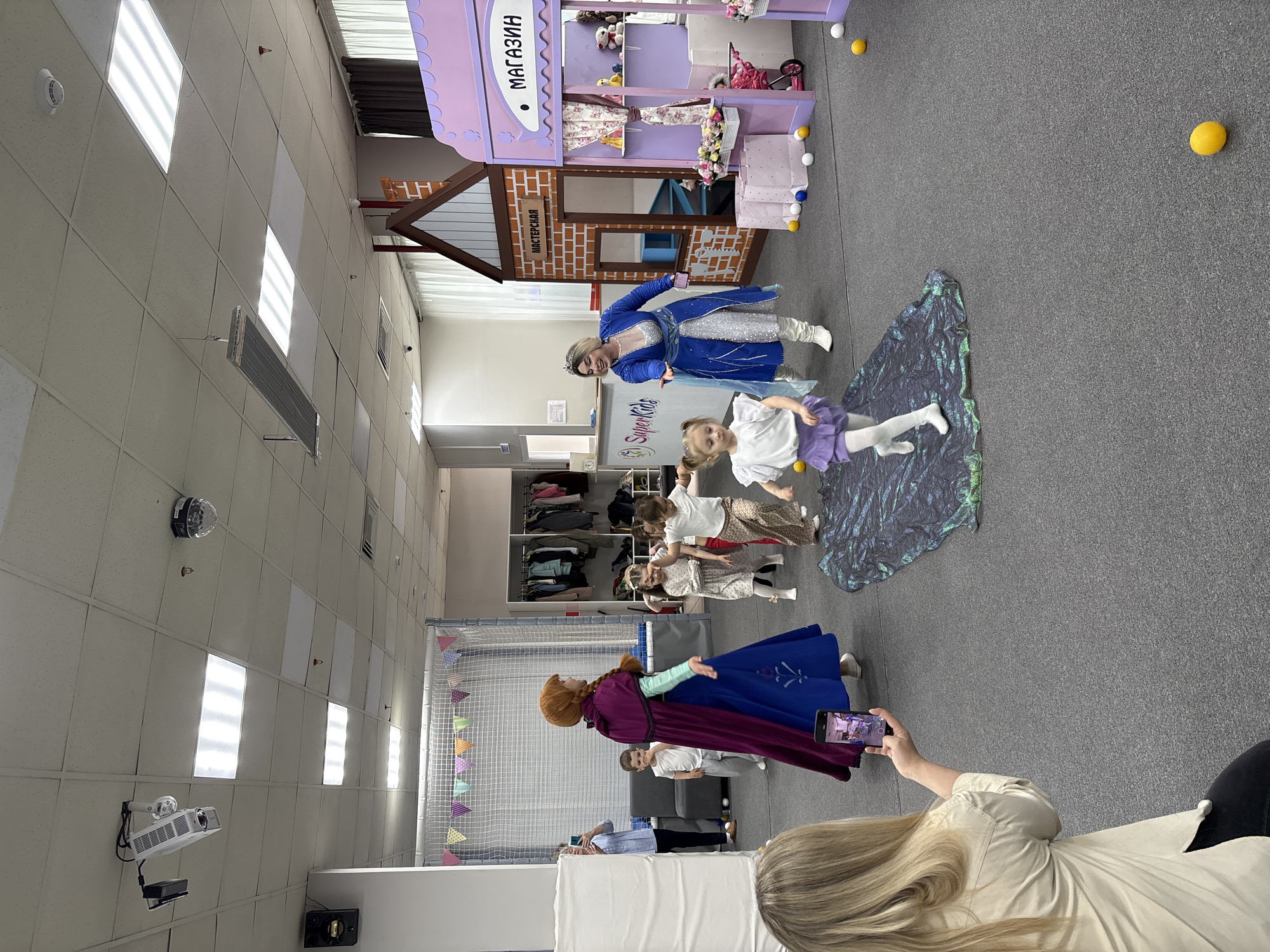 The image captures a scene inside a play area, likely in Krasnoyarsk, Russia given the Cyrillic writing on the playhouse. A woman dressed as Queen Elsa from Frozen stands next to a playhouse with the word "МАГАЗИН" (store) displayed. Another woman dressed as Princess Anna from Frozen is on the opposite side of the image. Several children are interacting with the characters. One child stands on a shimmering blue and green fabric, appearing to be dancing. A woman with blonde hair is holding a phone to record the scene. The environment includes a white ceiling with lighting fixtures, a grey carpeted floor, and various decorations, including colorful pennants hanging from a white net enclosure. White shelving units containing clothing can be seen in the background. Yellow and white balls are scattered on the floor.