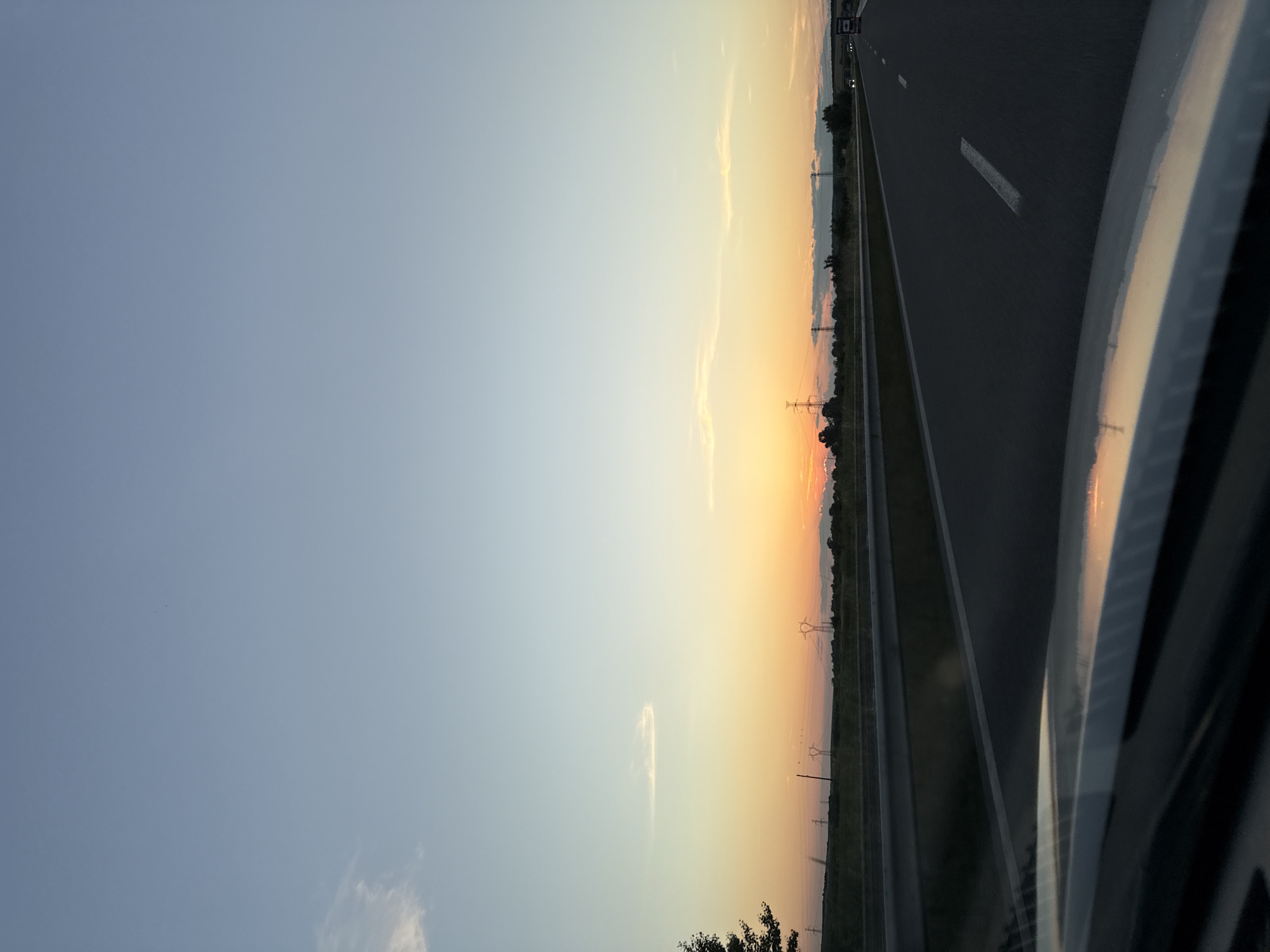 This image captures a sunset over a highway, likely in a rural or suburban area given the open fields and distant power lines. The sky transitions from a pale blue at the top to warm hues of orange and yellow near the horizon, indicating the sun is setting. A few thin clouds are visible. The highway itself is dark asphalt with white lane markings. A metal barrier separates the highway from the adjacent land. In the distance, several tall power pylons are silhouetted against the sunset. On the right side of the highway, a few vehicles, including a truck, are visible. The foreground shows a reflection on the hood of a car, possibly the camera's vehicle, showing the sunset colors. The overall scene evokes a sense of travel and the end of the day.
