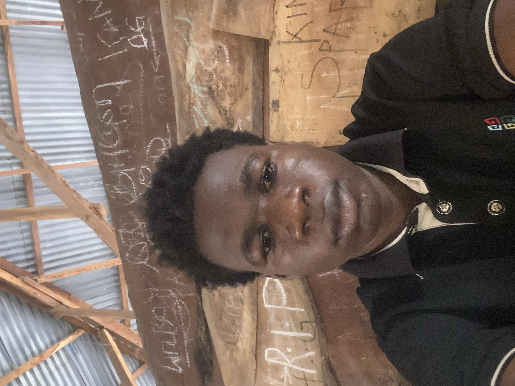 A dark-skinned person is captured in a close-up selfie indoors. The individual has short, dark, curly hair and appears slightly shiny or sweaty. They are wearing a dark polo shirt with a white stripe on the collar and a multicolored emblem on the left chest. The background consists of wooden beams and corrugated metal roofing, suggesting an informal or unfinished structure. Graffiti-like markings are visible on the wooden beams. While the context suggests the location might be Mararaba, Nigeria due to prior information, this cannot be definitively determined from the image alone.