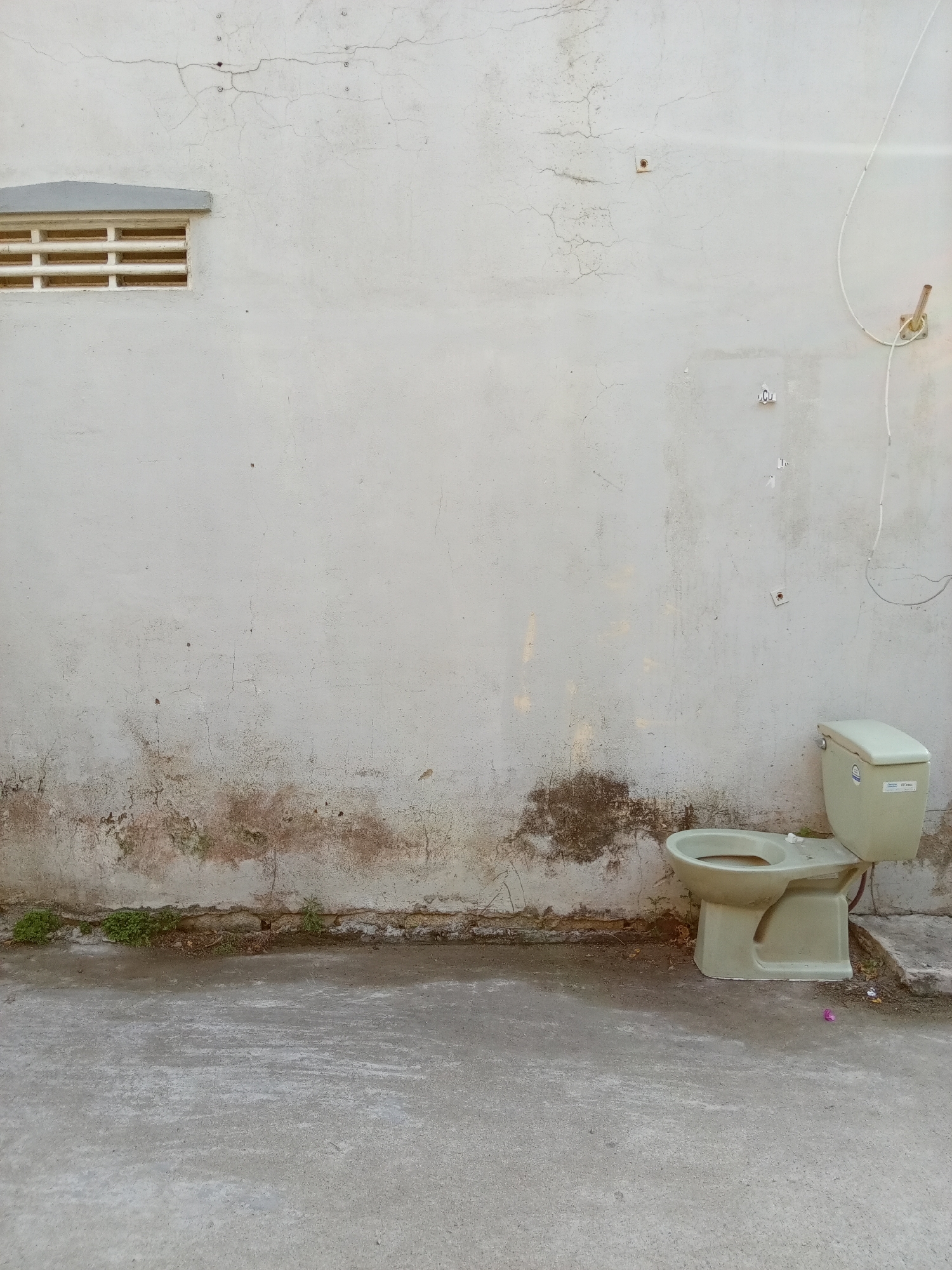 A discarded, light green toilet stands against a weathered white wall in an outdoor setting. The wall shows signs of age, with patches of discoloration and cracked paint. A small vent is visible near the top of the wall, and exposed wiring hangs down on the right. The ground is a concrete surface. The location is Bắc Nha Trang, Vietnam.
