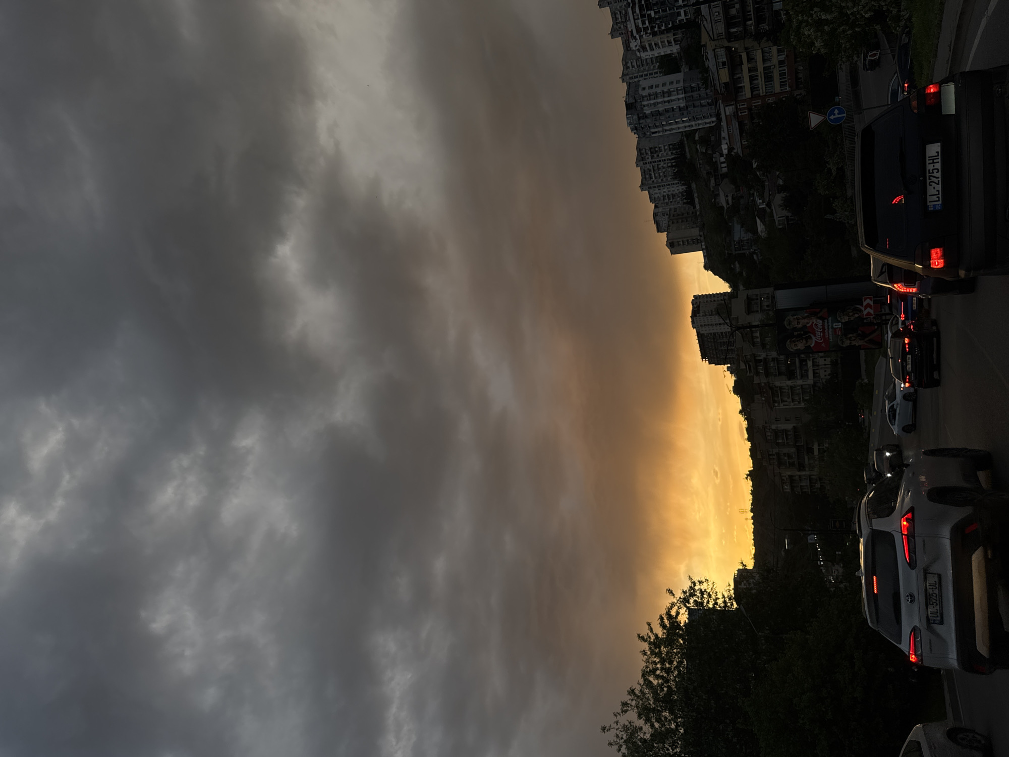 The image depicts a street scene in Tbilisi, Georgia, during a dramatic sunset or approaching storm. The sky transitions from dark, ominous clouds on the left to a bright, golden hue on the right. Buildings are visible in the background, arranged on a hillside. A line of vehicles is on the road, including a silver Toyota with the license plate "LL-552B-UL" and a black car with the license plate "LL-275-HL". Trees are in the foreground, silhouetted against the bright sky. A traffic sign is present.